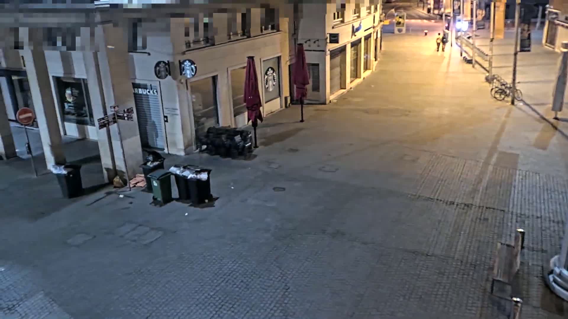 At night, an empty city street shows several trash cans near a closed Starbucks and two closed umbrellas.