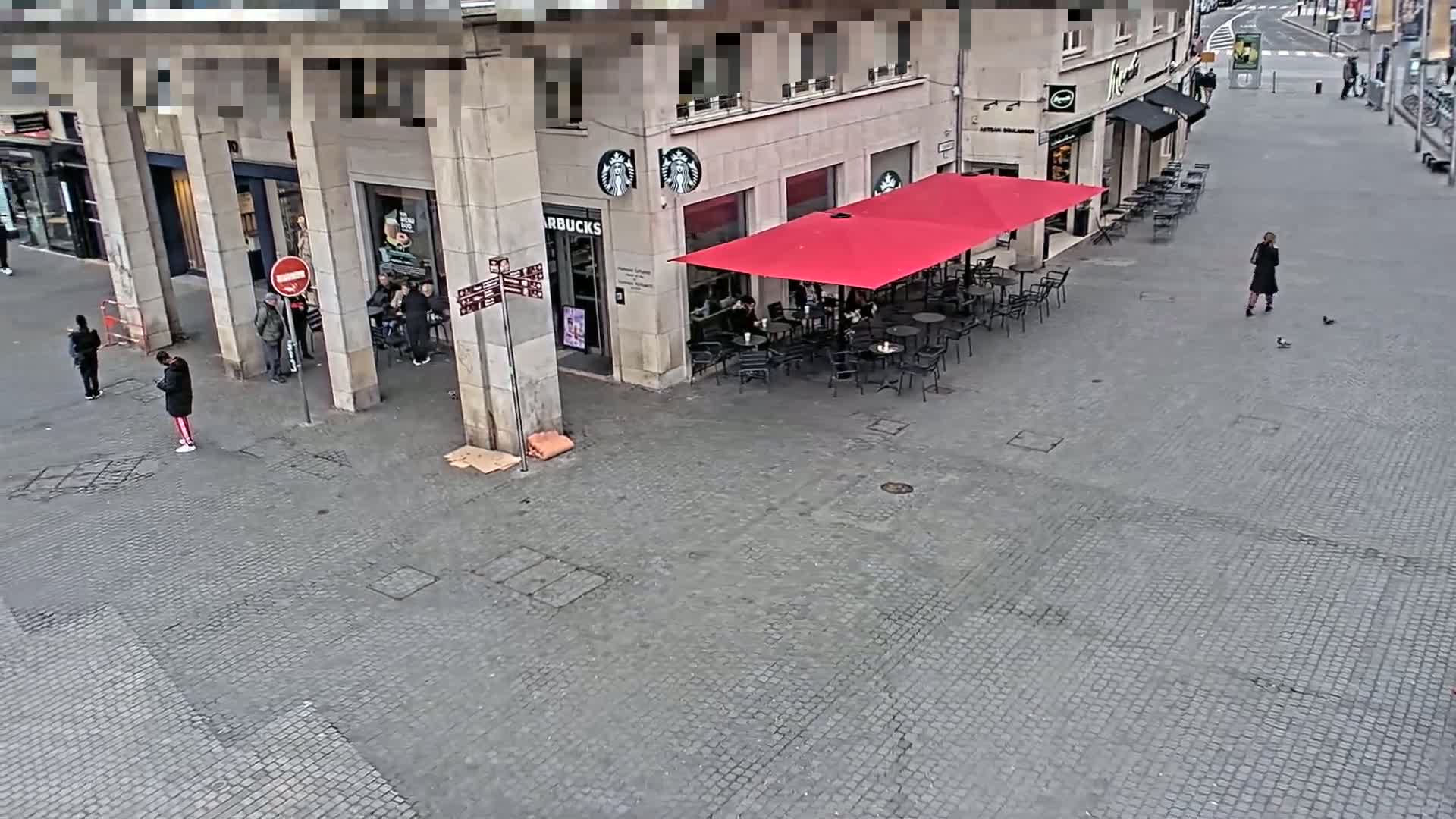 A paved outdoor area features a Starbucks with a red umbrella-covered patio, several people standing around, and a woman walking by.