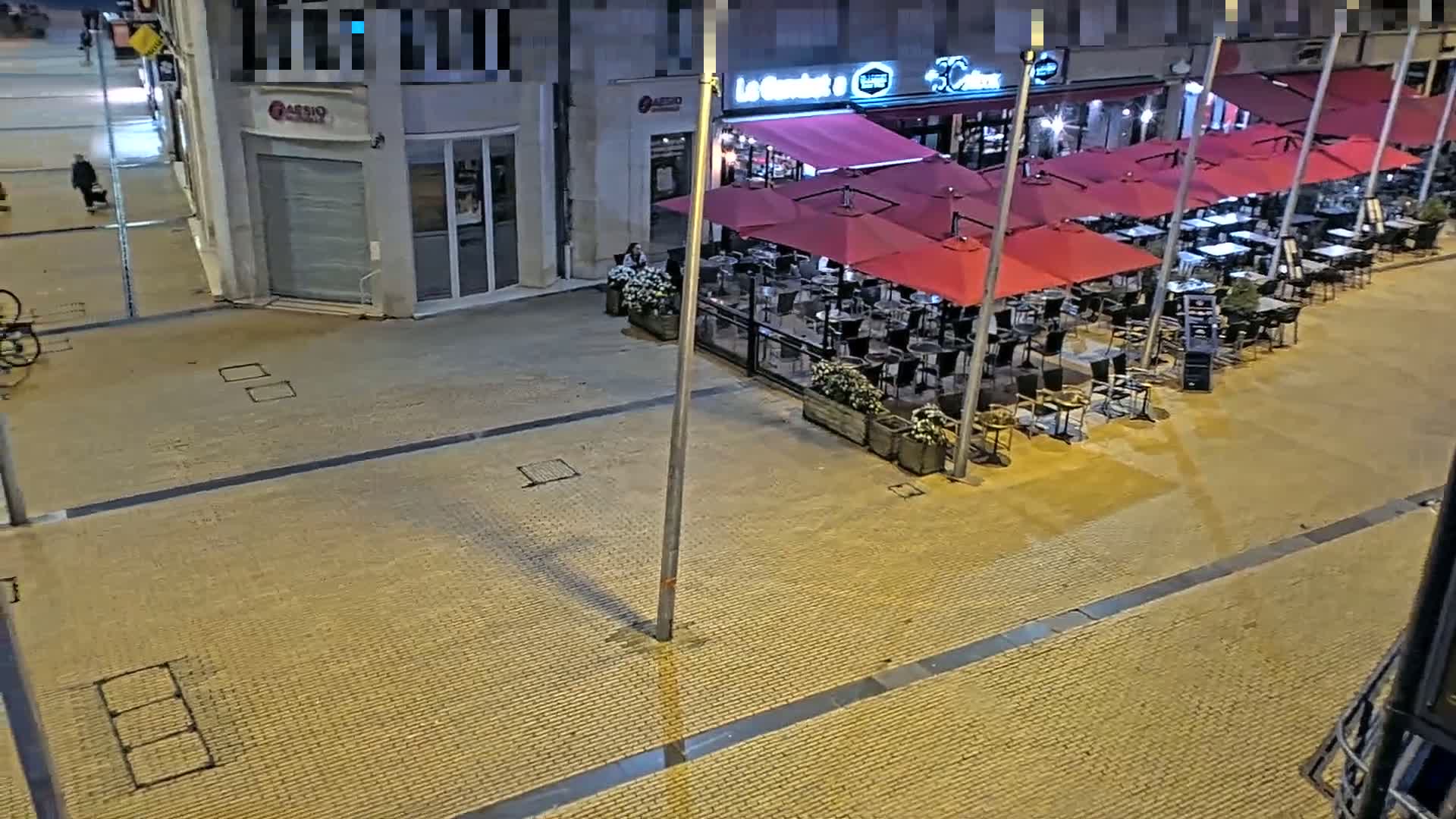An outdoor nighttime scene shows a mostly empty restaurant patio with numerous red umbrellas and metal tables and chairs, situated on a paved street.