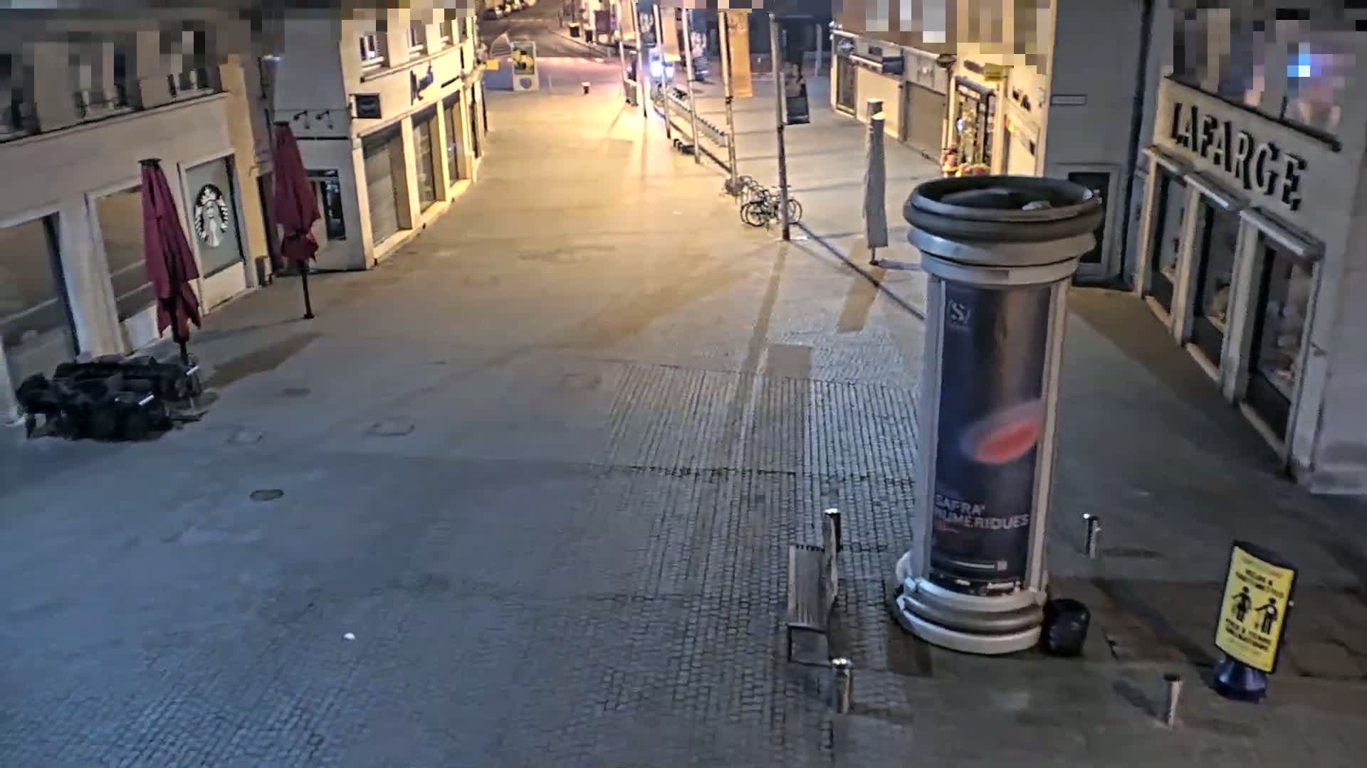 A mostly empty, paved street at night is lit by streetlights, with closed shops lining both sides and a few outdoor seating areas with closed umbrellas.