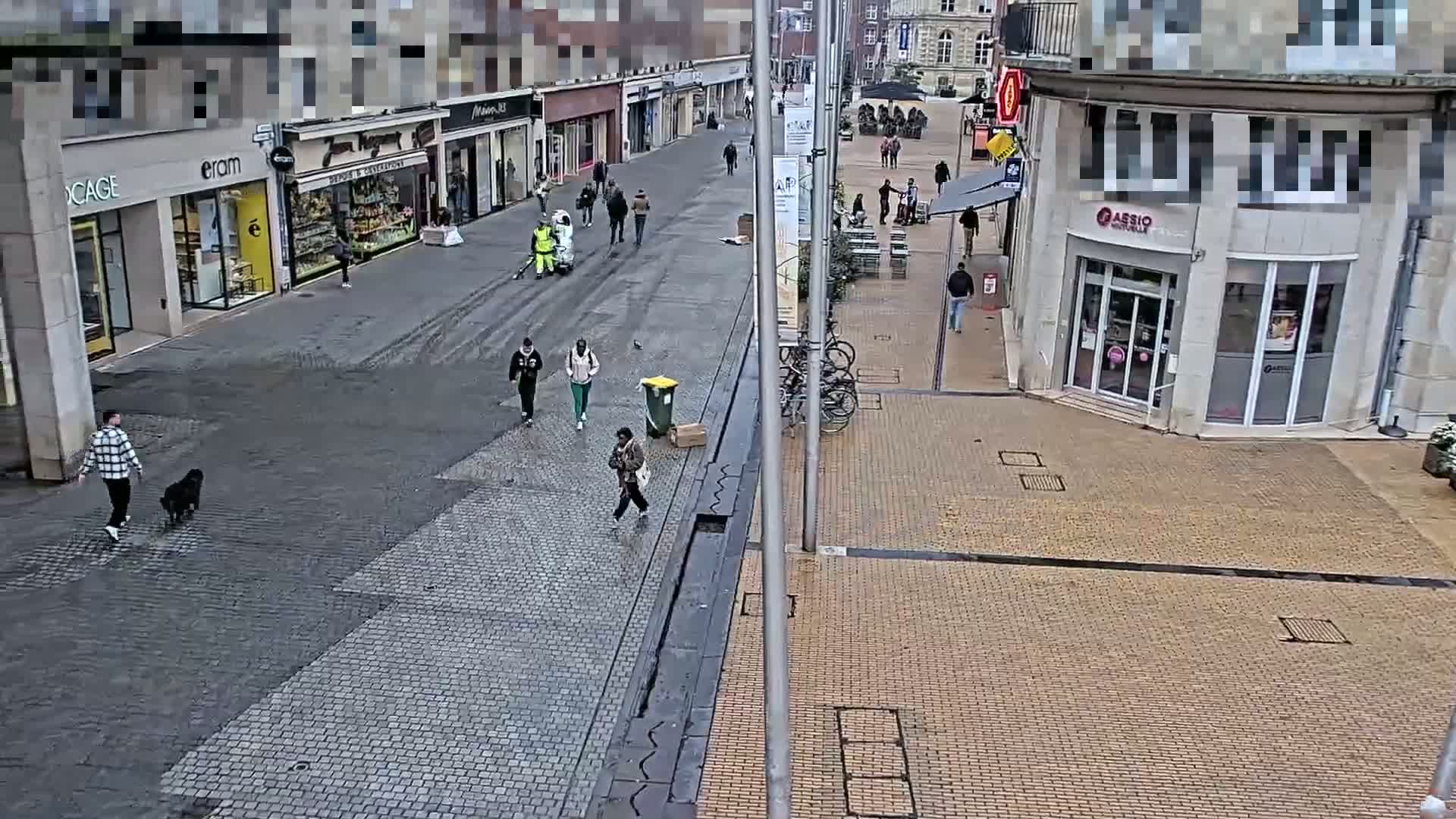 A mostly empty cobblestone street with several pedestrians and a person cleaning the street with a machine, under an overcast sky.