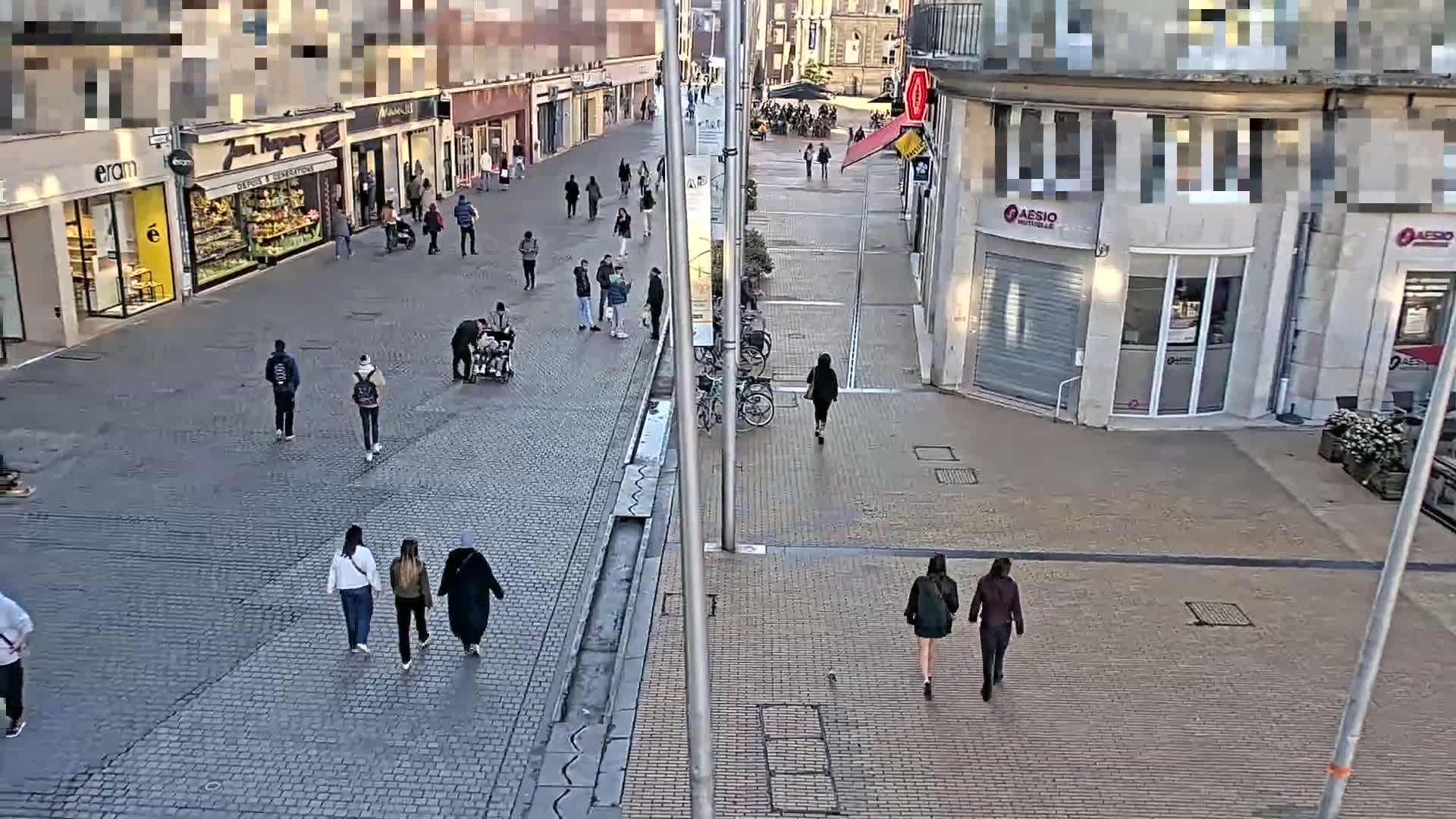 A mostly empty cobblestone pedestrian street with a few people walking past shops and businesses under a clear sky.
