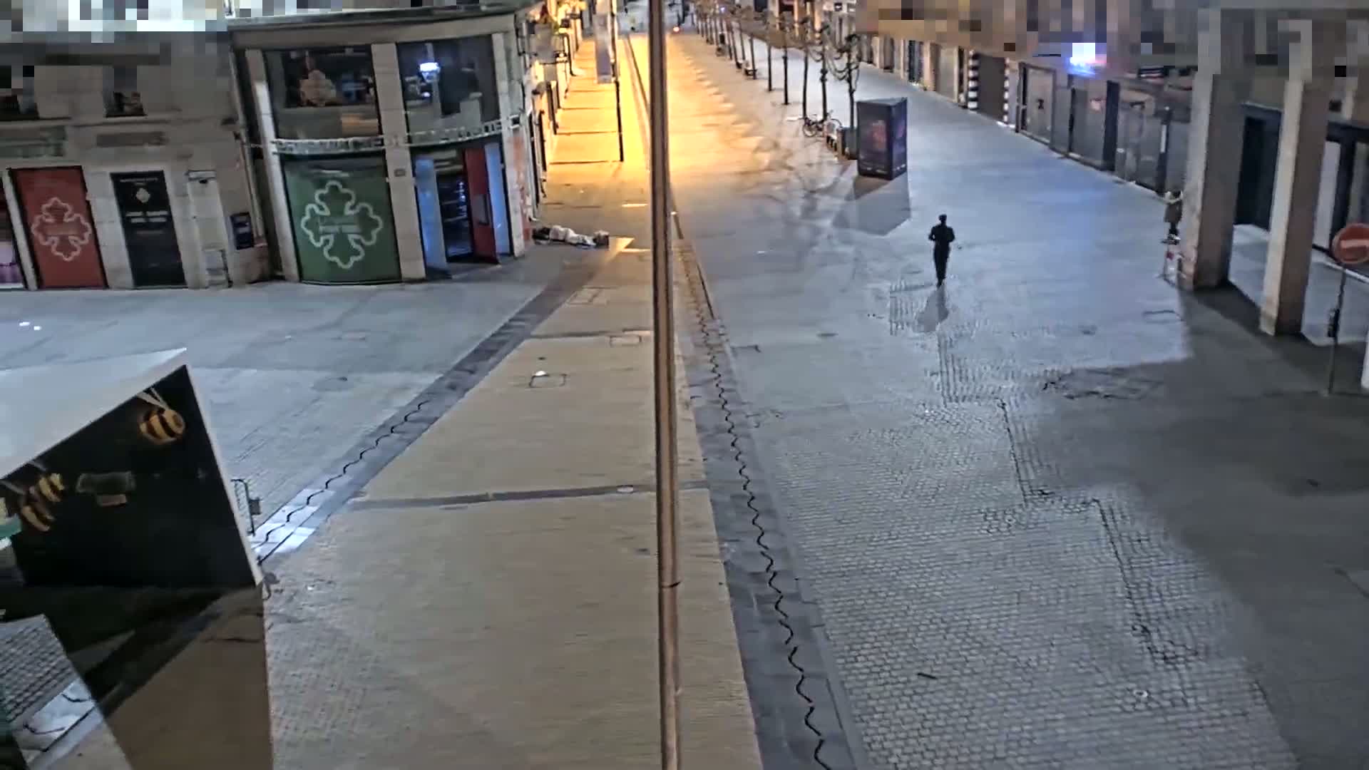 A person walks down a mostly empty, paved street at night, past a person lying on the ground near a building.