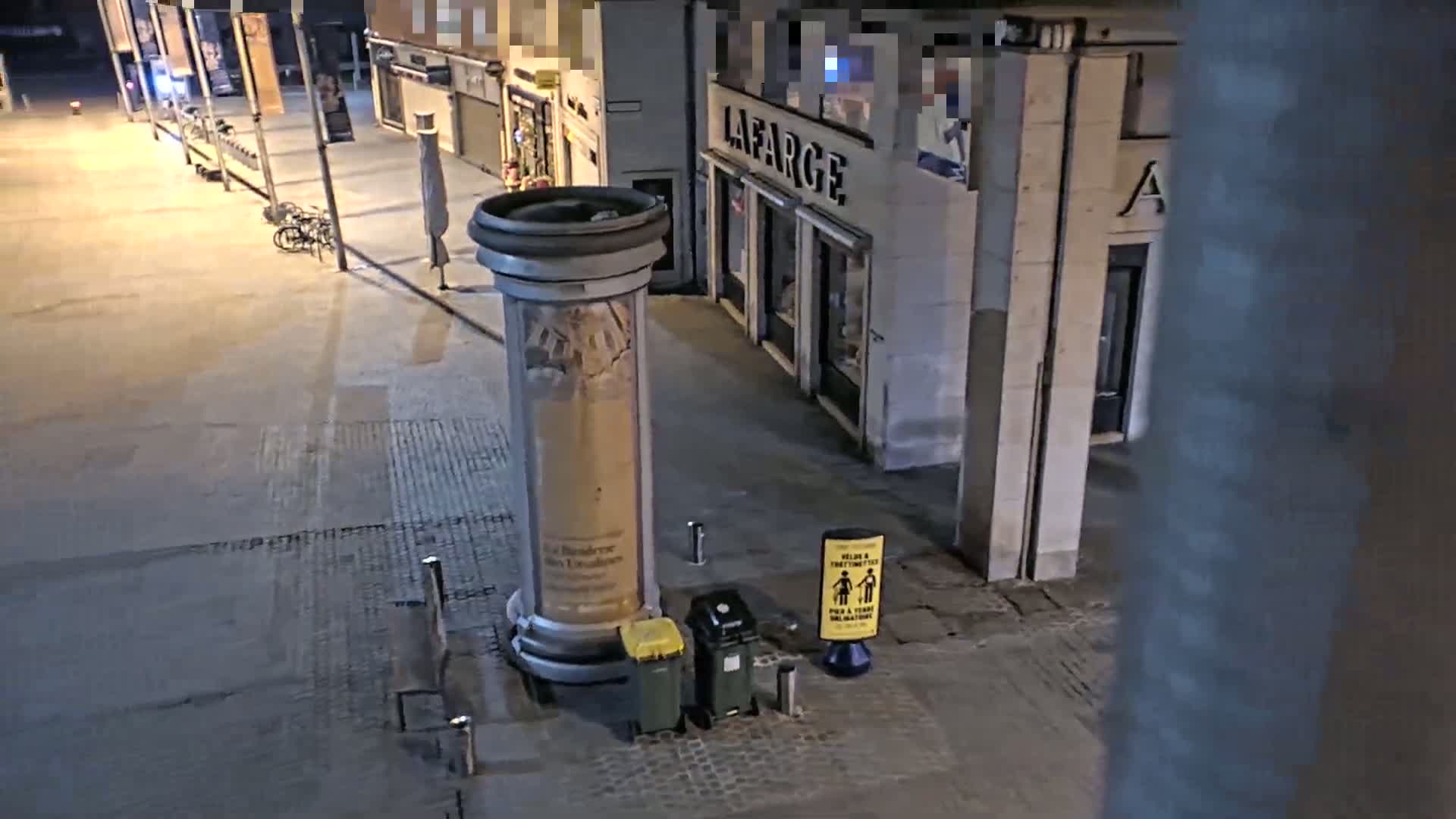 At night, an empty street scene shows a large advertising column next to a trash receptacle and a small yellow sign in front of closed shops.