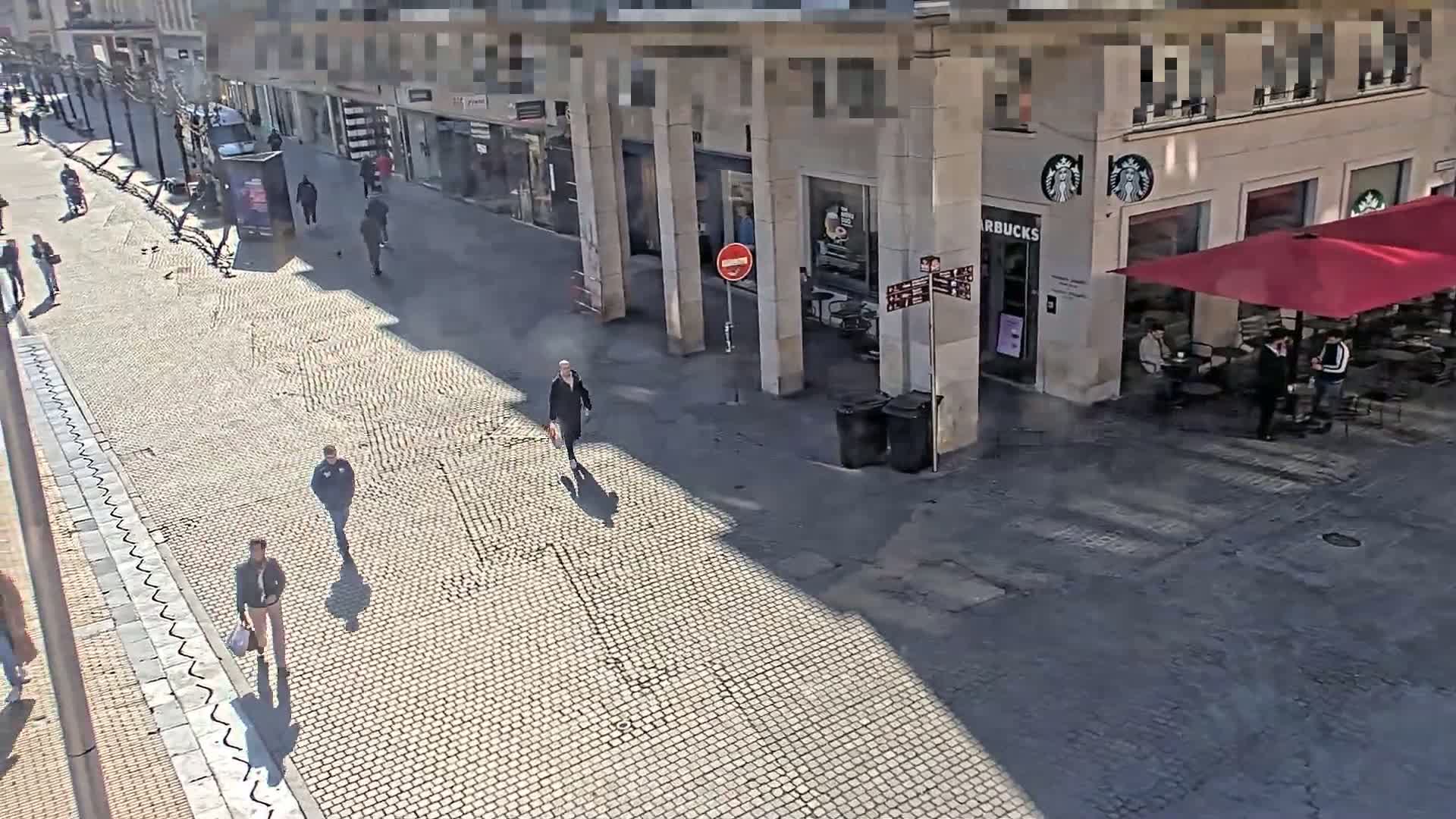 A cobblestone street scene on a sunny day shows several pedestrians walking past a Starbucks with outdoor seating under a red umbrella.