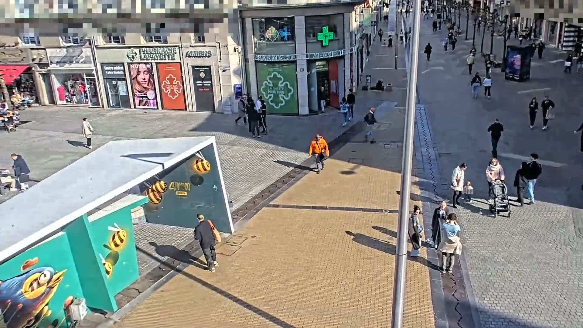 A sunny city square with pedestrians walking past shops and a modern structure featuring bee artwork.