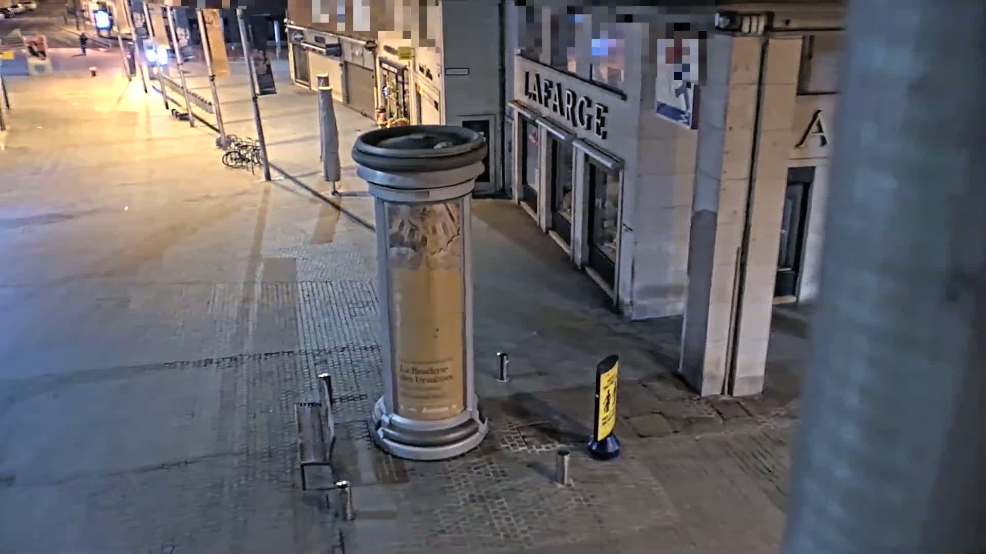A nighttime street scene shows an empty, paved pedestrian area with a large advertising column, a couple of benches, and a few parked bicycles near some storefronts.