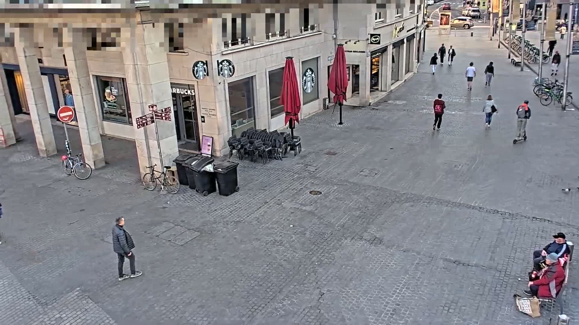 A cobblestone plaza with a few pedestrians, stacked chairs and trash cans in front of a Starbucks and other shops, under a mostly clear sky.