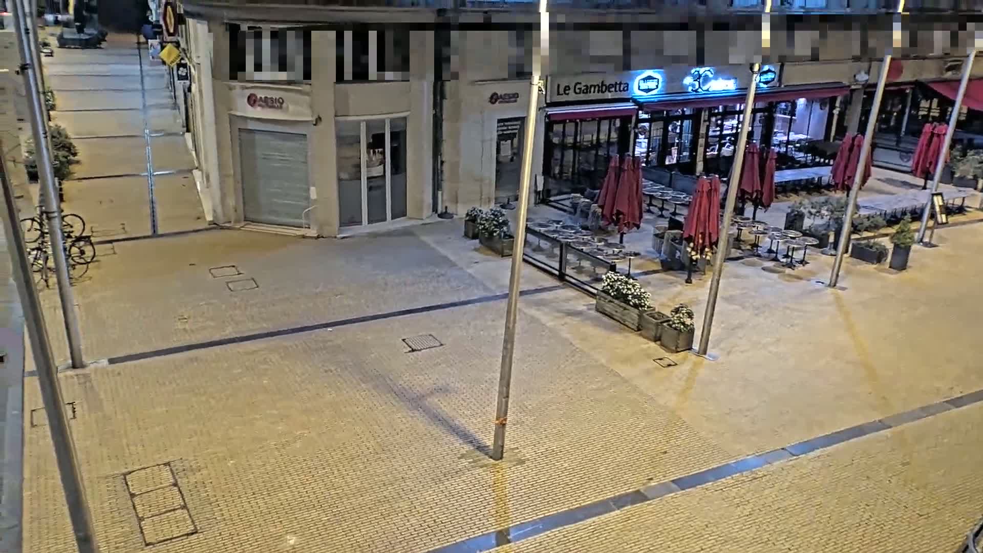 An empty, paved plaza at night with a restaurant's outdoor seating area featuring red umbrellas and several potted plants.