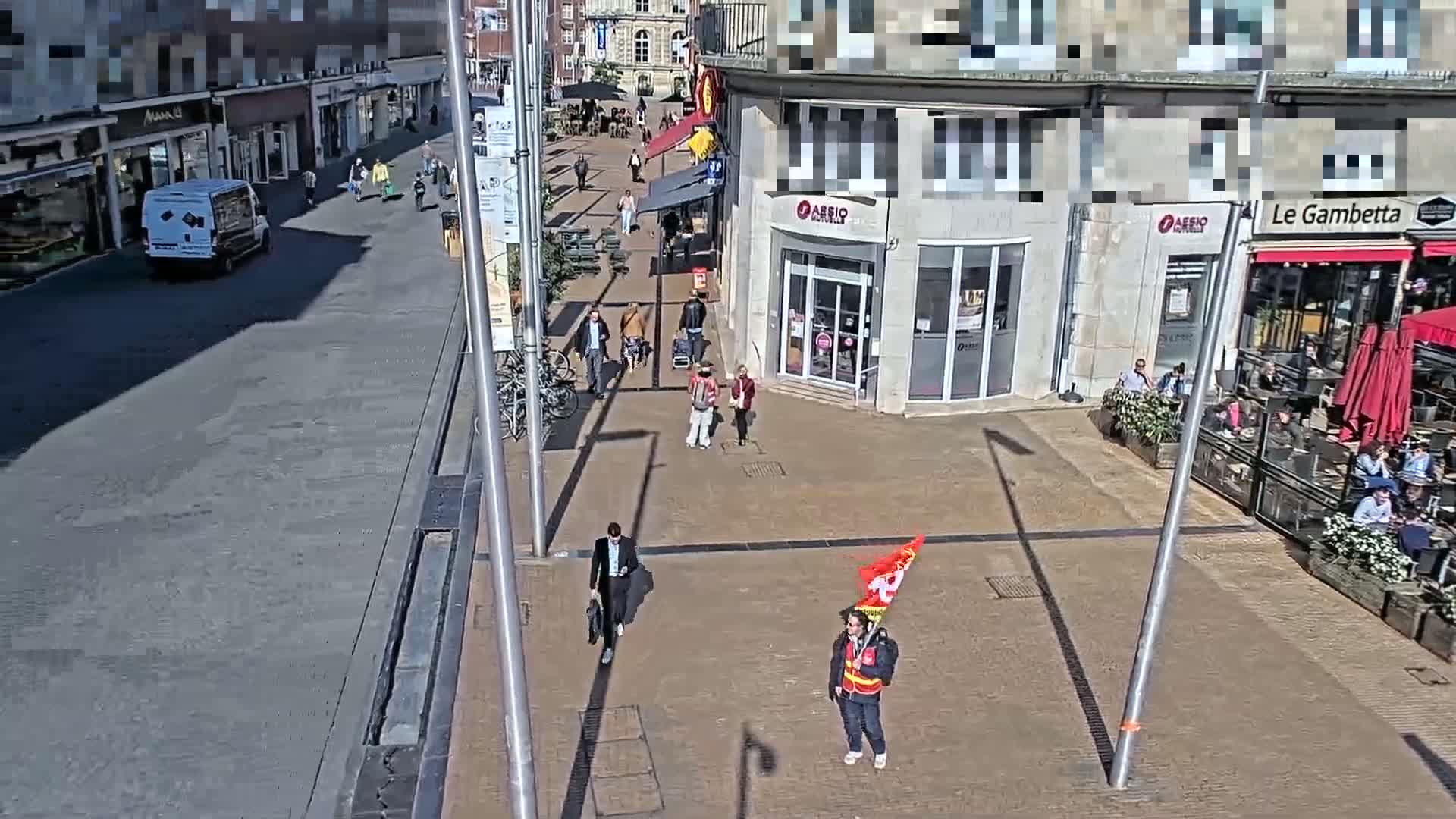 A sunny day on a paved pedestrian area with several people walking, a person holding a flag, and some people sitting at outdoor cafes.