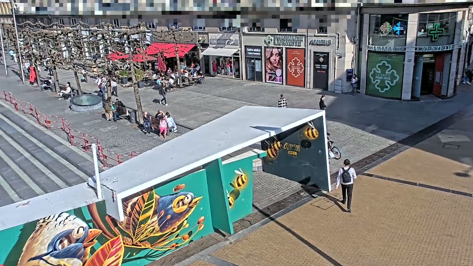 A sunny city square with several people walking around, sitting at outdoor cafes, and near a building with a mural of birds and bees.