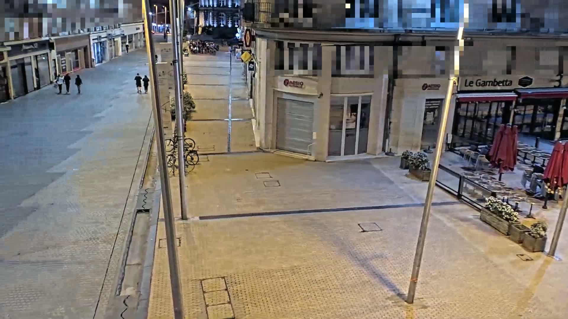 A mostly empty, paved street at night is shown, with a few pedestrians and bicycles visible, and an outdoor restaurant patio with closed umbrellas.