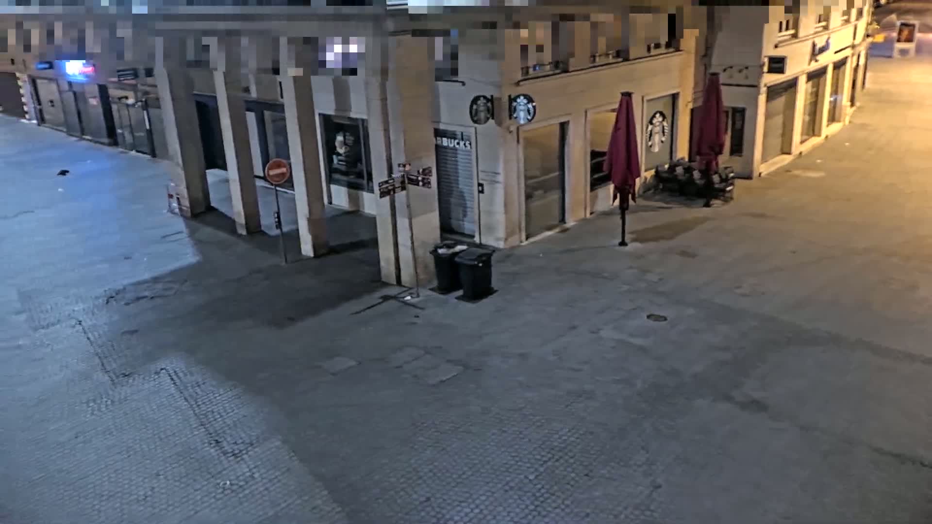 An empty city street at night, showing a Starbucks with closed shutters, outdoor seating, and two garbage cans.