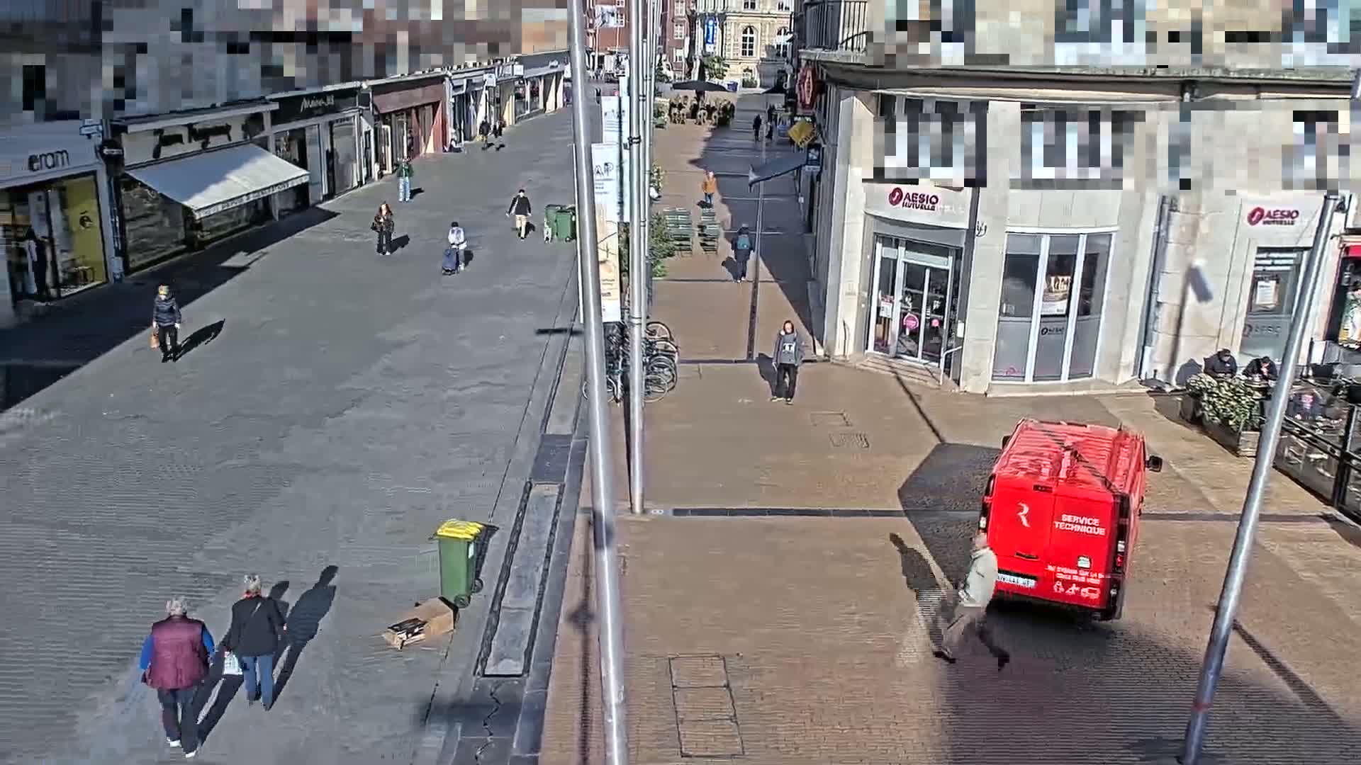 A mostly empty city street on a sunny day, with pedestrians walking and a red van parked near a building.
