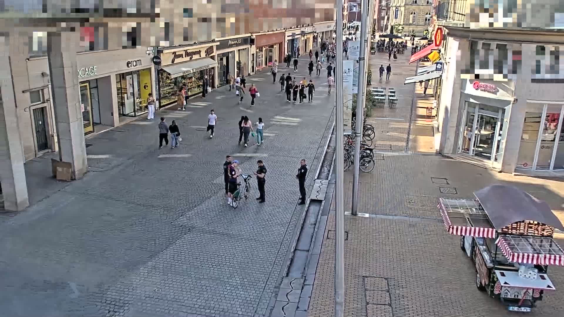 A paved pedestrian shopping street with several people walking, some police officers talking to a group of people near a bicycle, and a food cart parked on the side on a sunny day.