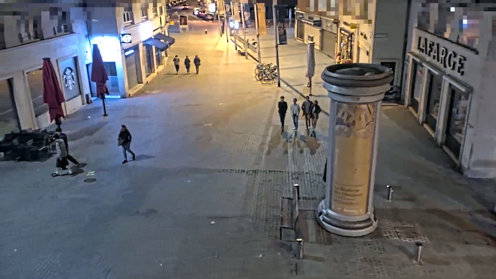 At night, several people walk on a paved street lined with buildings, under dim lighting.