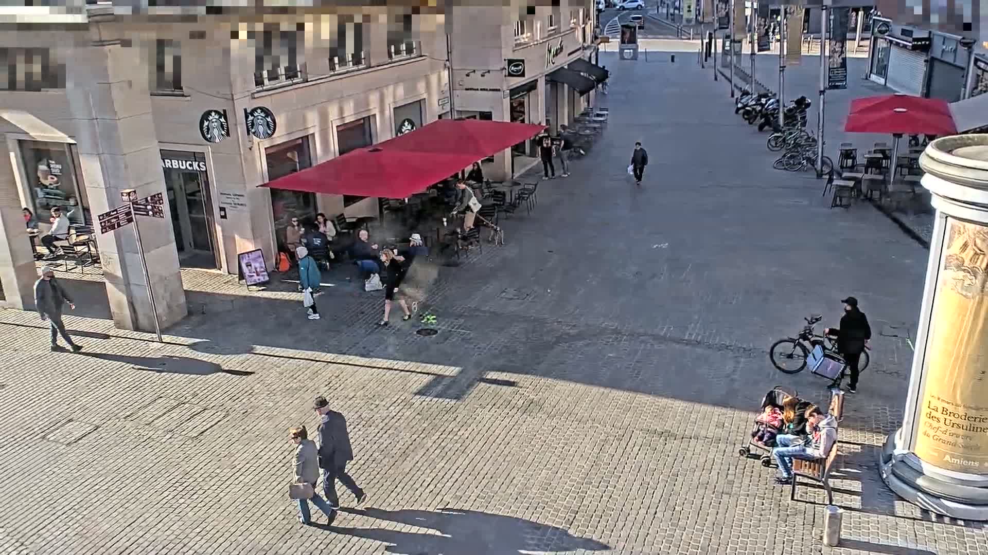 A sunny city square features a Starbucks, outdoor seating areas with red umbrellas, pedestrians, and a person on a bicycle; several people are sitting on a bench.