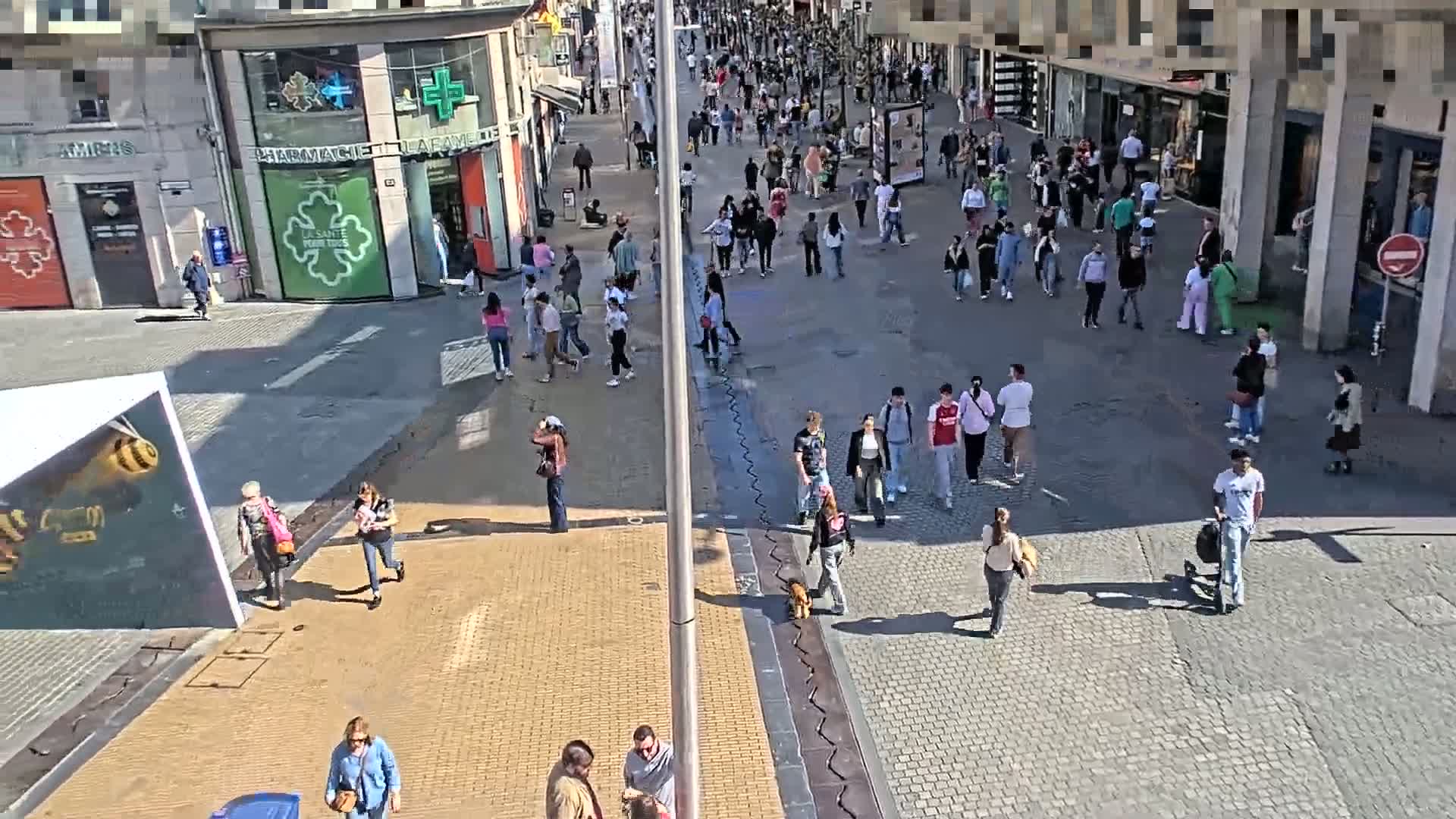 A sunny day reveals a bustling pedestrian street with many people walking in various directions, passing shops and buildings.