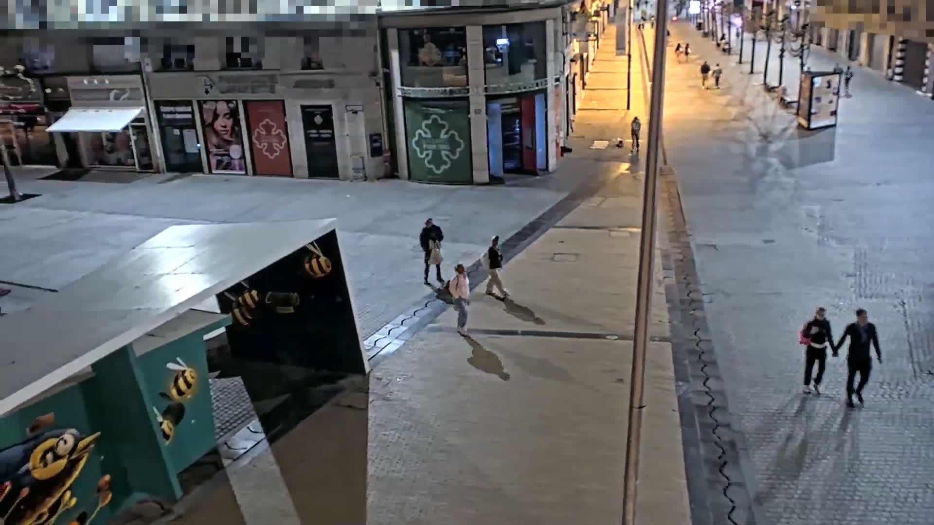 A nighttime street scene shows several pedestrians walking on a paved area near buildings, with some people near a structure decorated with bee images.