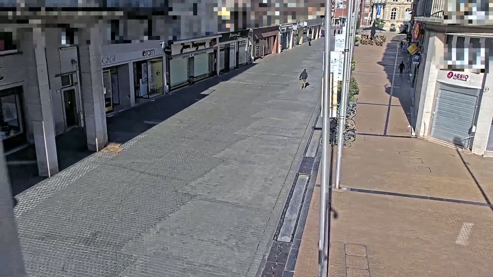 A mostly empty cobblestone street lined with shops is seen on a sunny day, with a single person walking down the center.