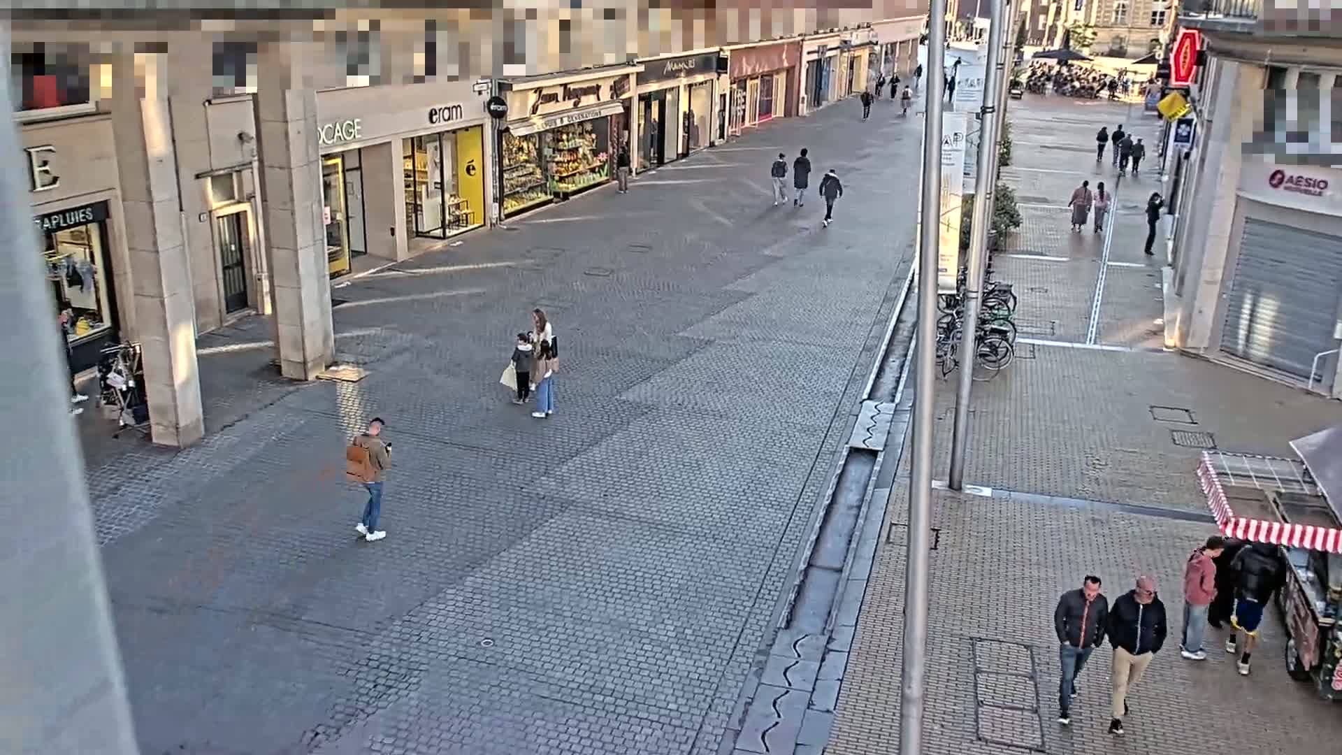A mostly empty cobblestone pedestrian street lined with shops is seen on a sunny day, with a few people walking and standing in small groups.