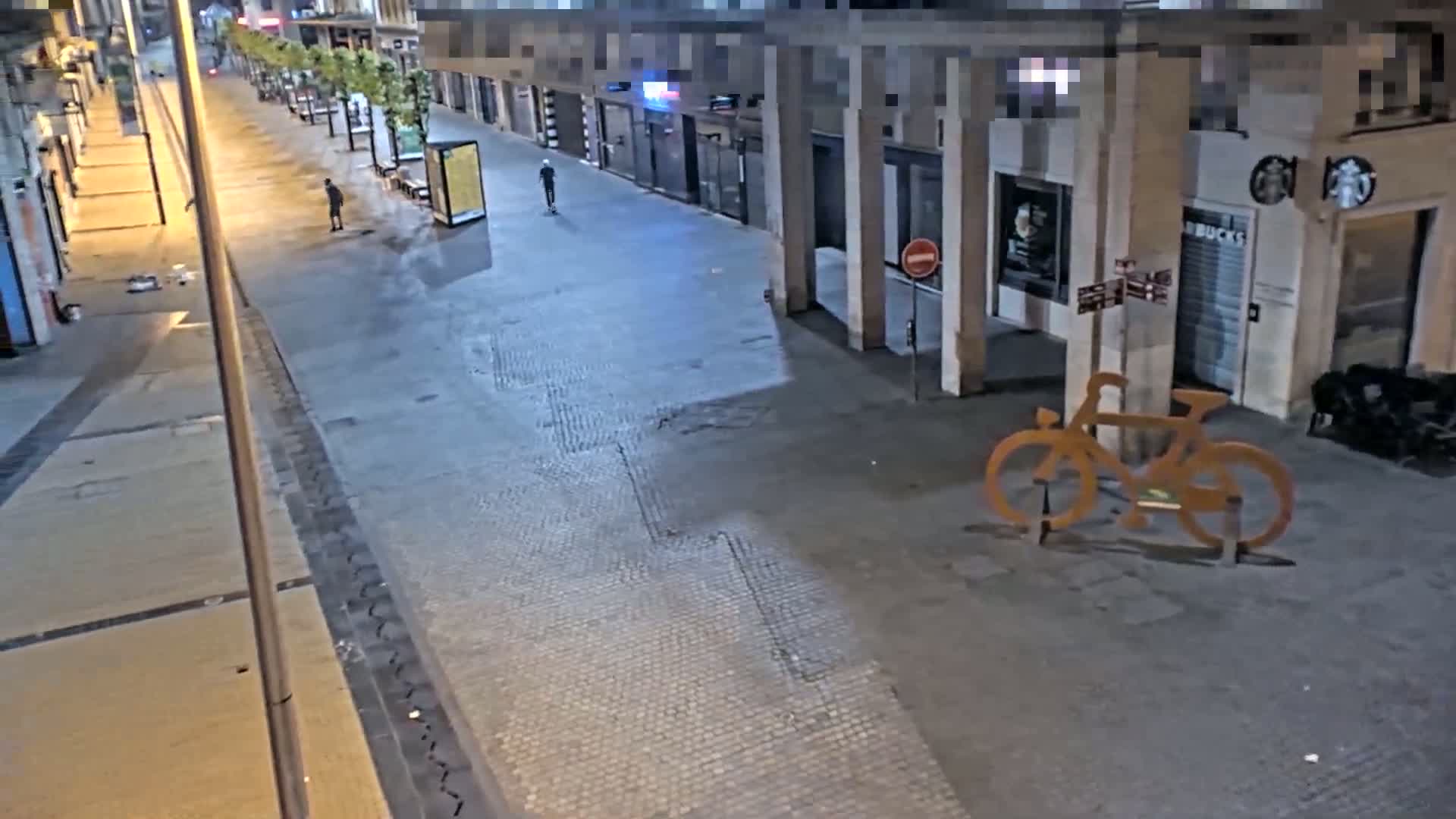 An empty city street at night is illuminated by streetlights, showing a few pedestrians and a large metal bicycle sculpture.