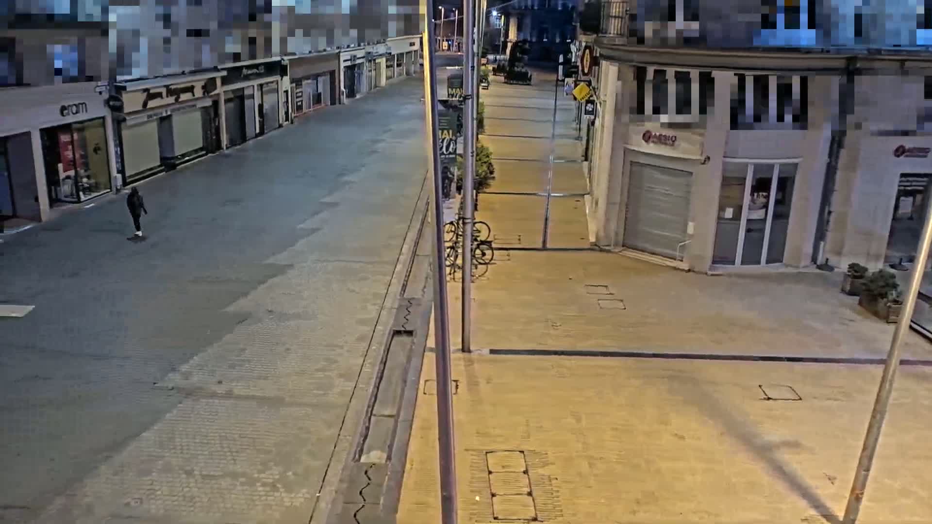 A person walks down a mostly empty street at night, past some parked bicycles and closed shops.