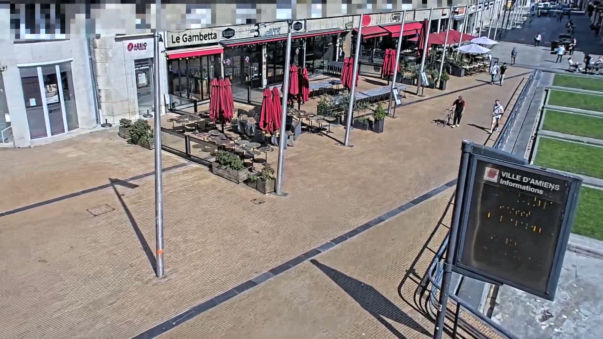 A sunny outdoor cafe scene with red umbrellas and several people walking by.