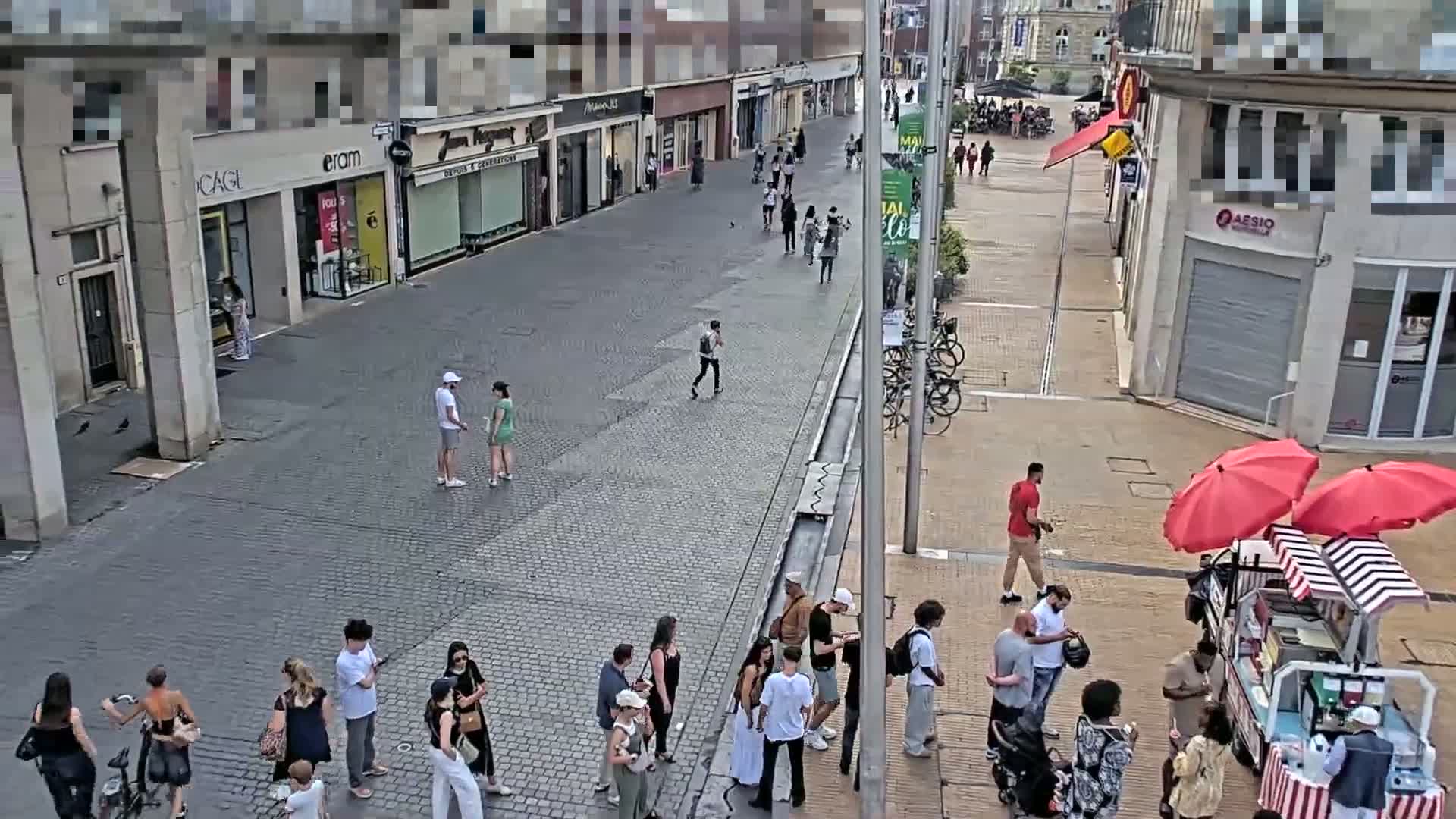 A paved pedestrian street with shops and people walking on a sunny day, some people are standing in lines near a food cart with red umbrellas.