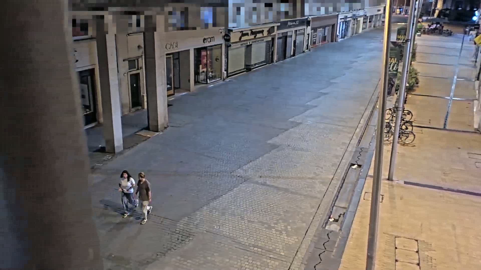 Two people walk down an empty, paved street at night, past closed shops under a nighttime sky.