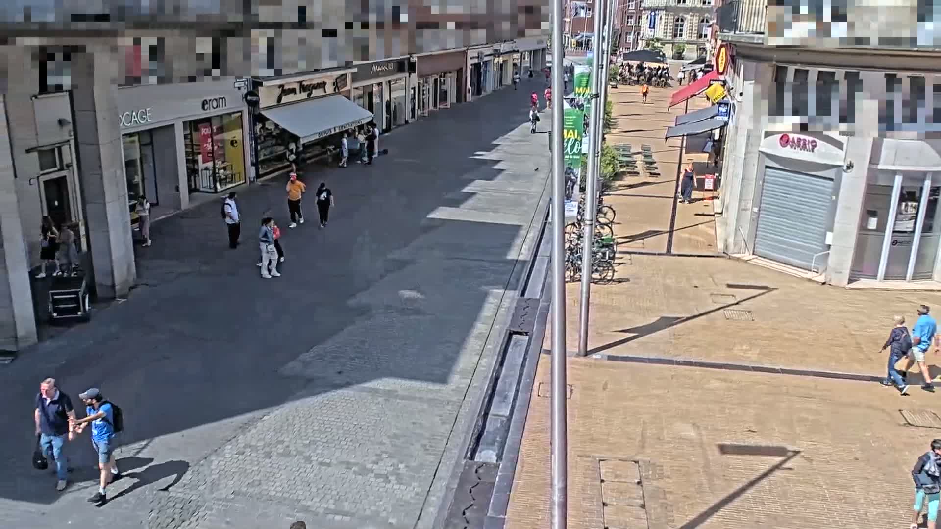 A sunny city street scene shows several pedestrians walking past shops and parked bicycles.