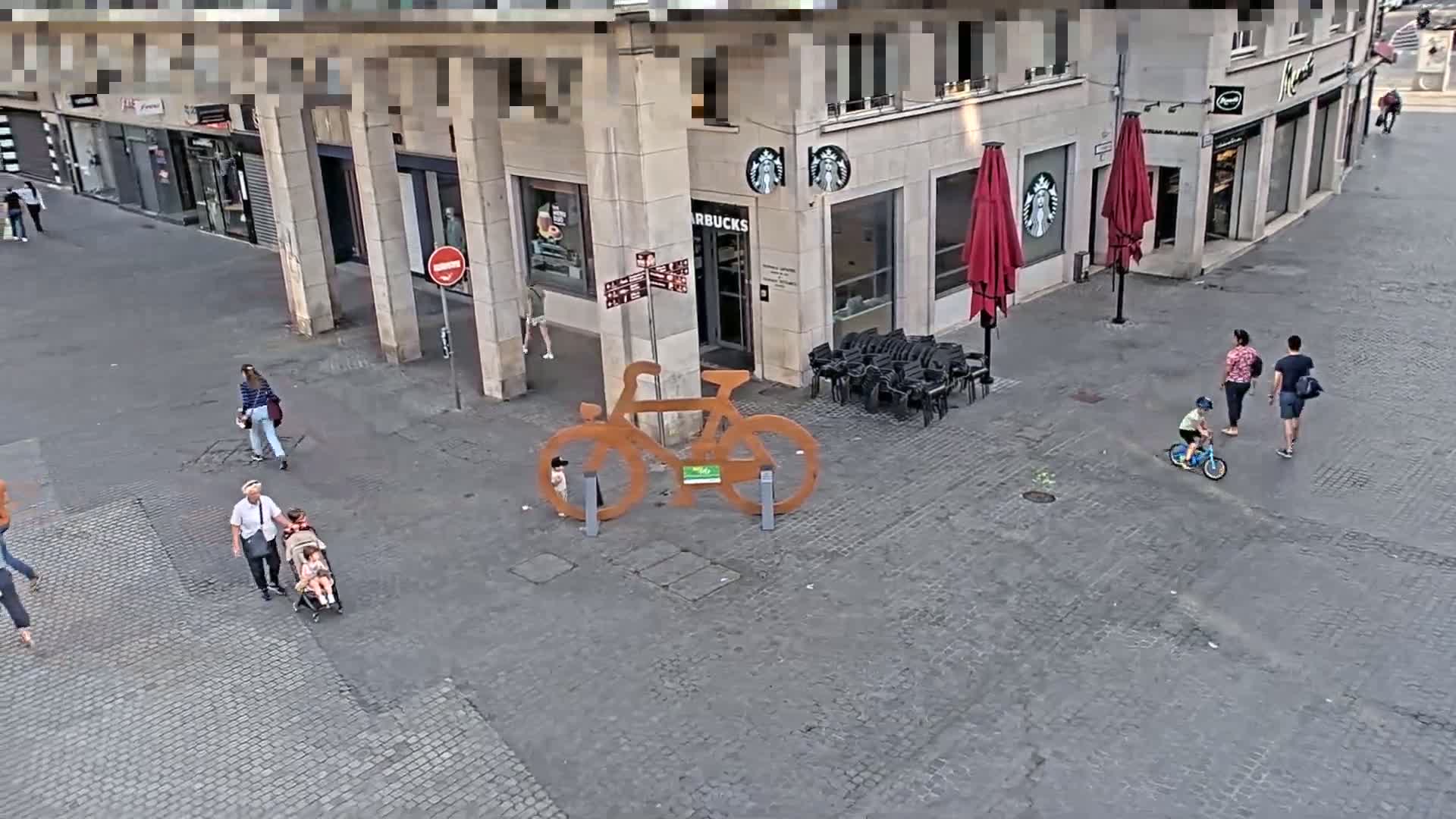 A large metal bicycle sculpture stands in a cobblestone city square where several people walk, and a small child rides a bicycle.