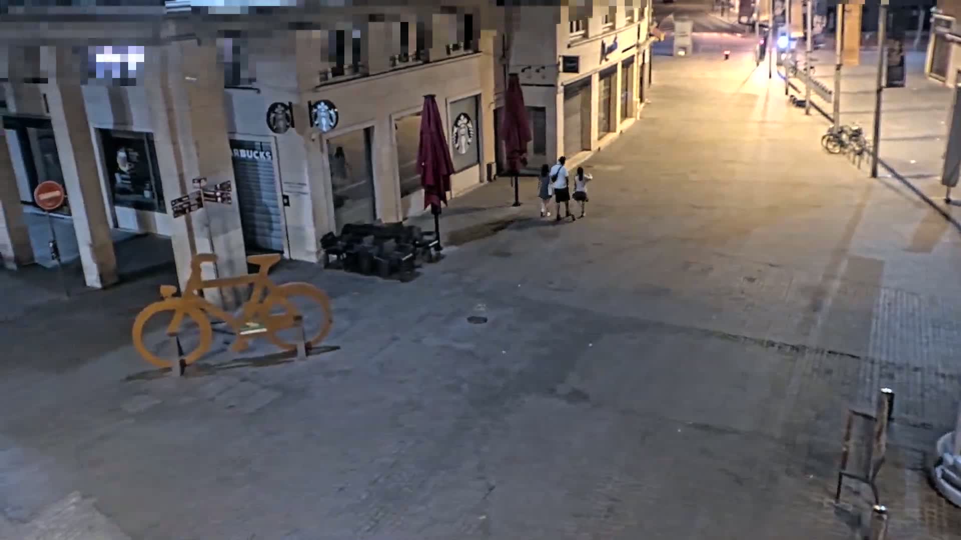 A nighttime, outdoor view shows a mostly empty street with three people walking past a Starbucks, a large metal bicycle sculpture, and a parked bicycle.