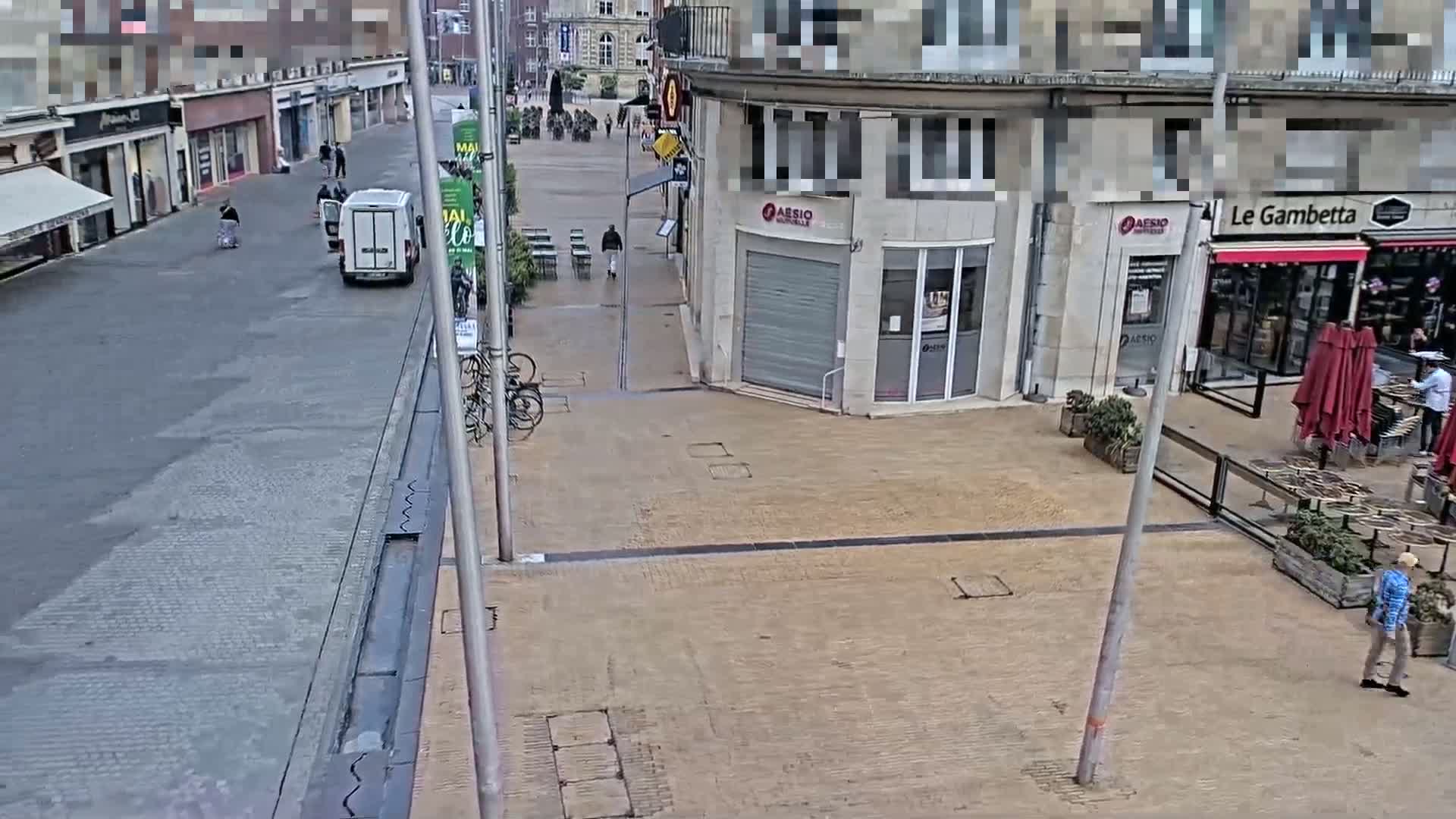 A mostly empty town square with a few pedestrians and a white van, several bicycles leaning against a pole, and outdoor cafe seating.