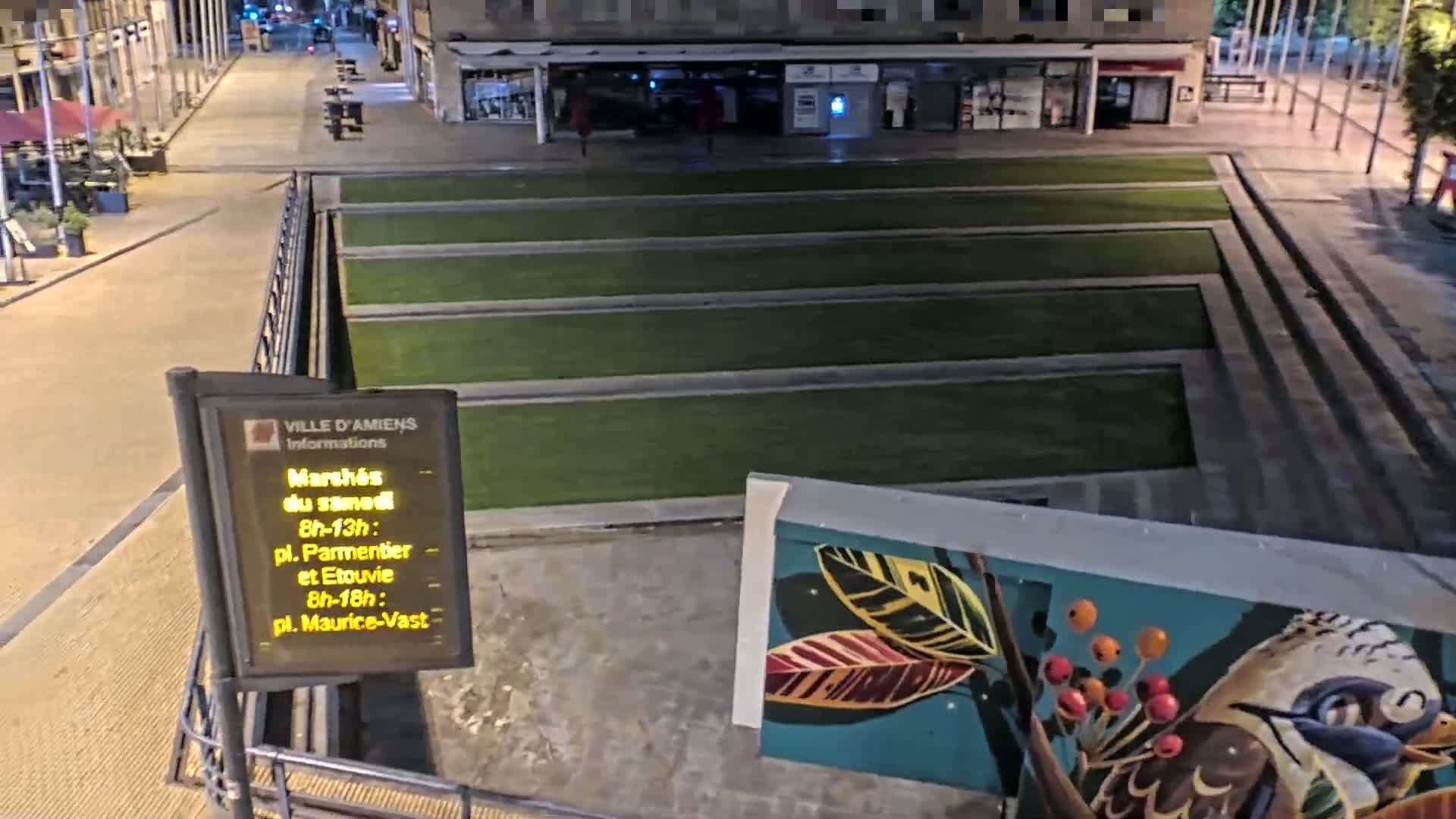 A nighttime view of a paved plaza with rectangular sections of artificial turf, a mural featuring birds and leaves, and a digital sign.
