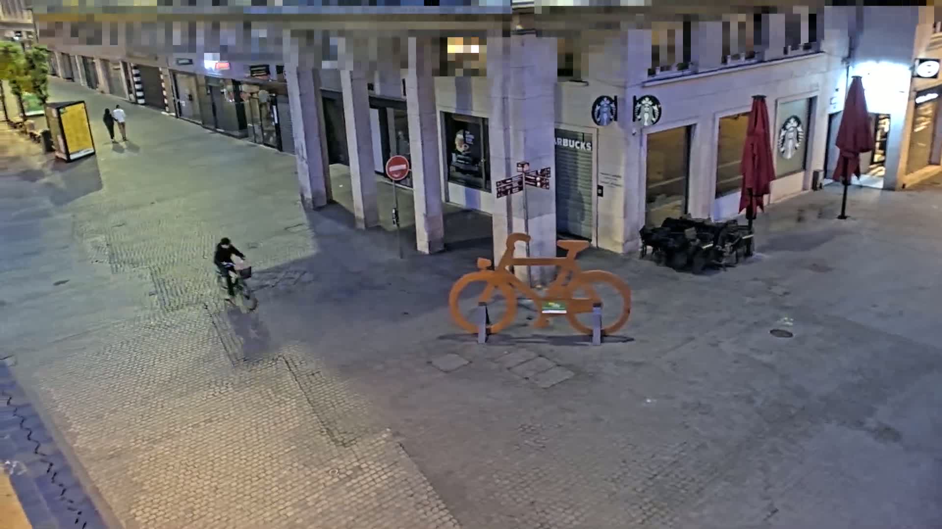 A person rides a bicycle on a cobblestone street at night past a Starbucks and a large metal bicycle sculpture.