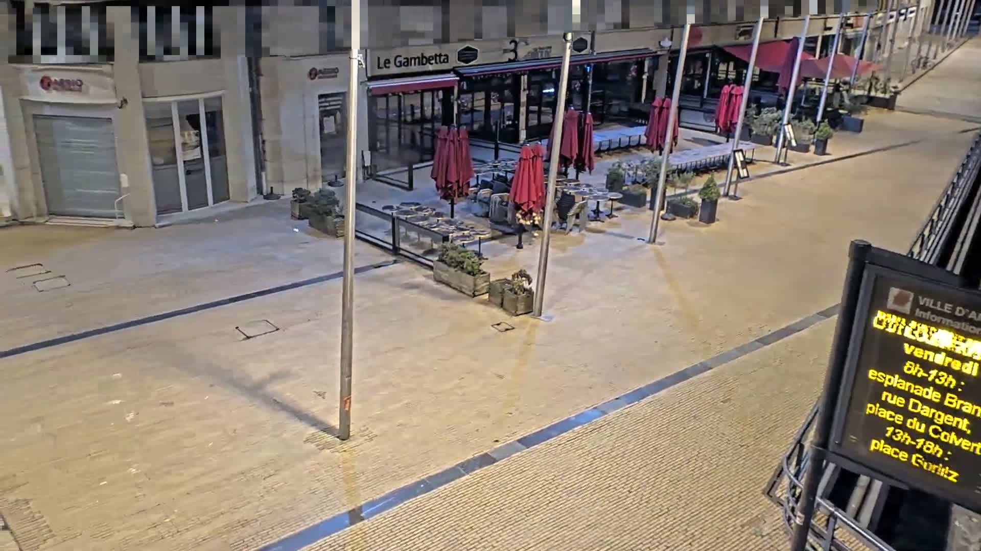 An outdoor nighttime scene shows an empty plaza with several closed restaurants, some with outdoor seating areas containing closed red umbrellas.