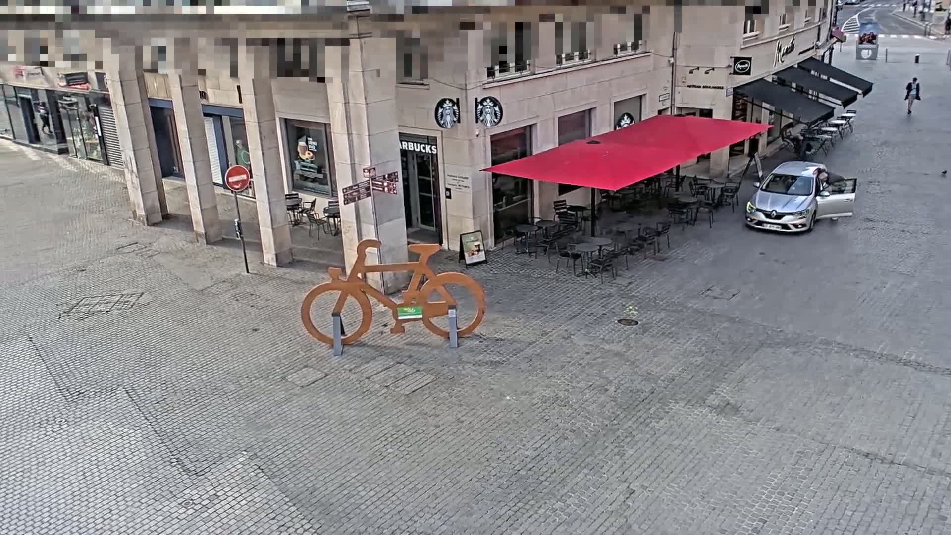A large, wooden bicycle sculpture stands in a cobblestone plaza next to a Starbucks with outdoor seating under a red awning, and a silver car is parked nearby.