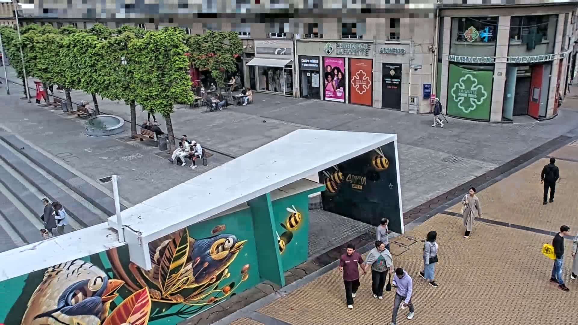 A paved town square with several people walking and sitting, featuring a mural of birds and bees on a modern structure, under a partly cloudy sky.