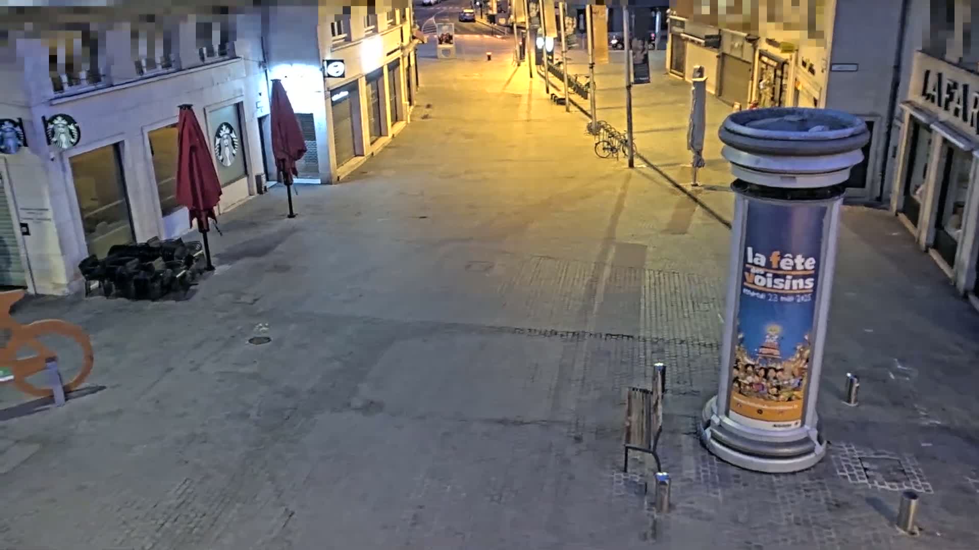 An empty city street at night is illuminated by streetlights, showing closed shops, a few parked bicycles, and a large advertisement on a column.
