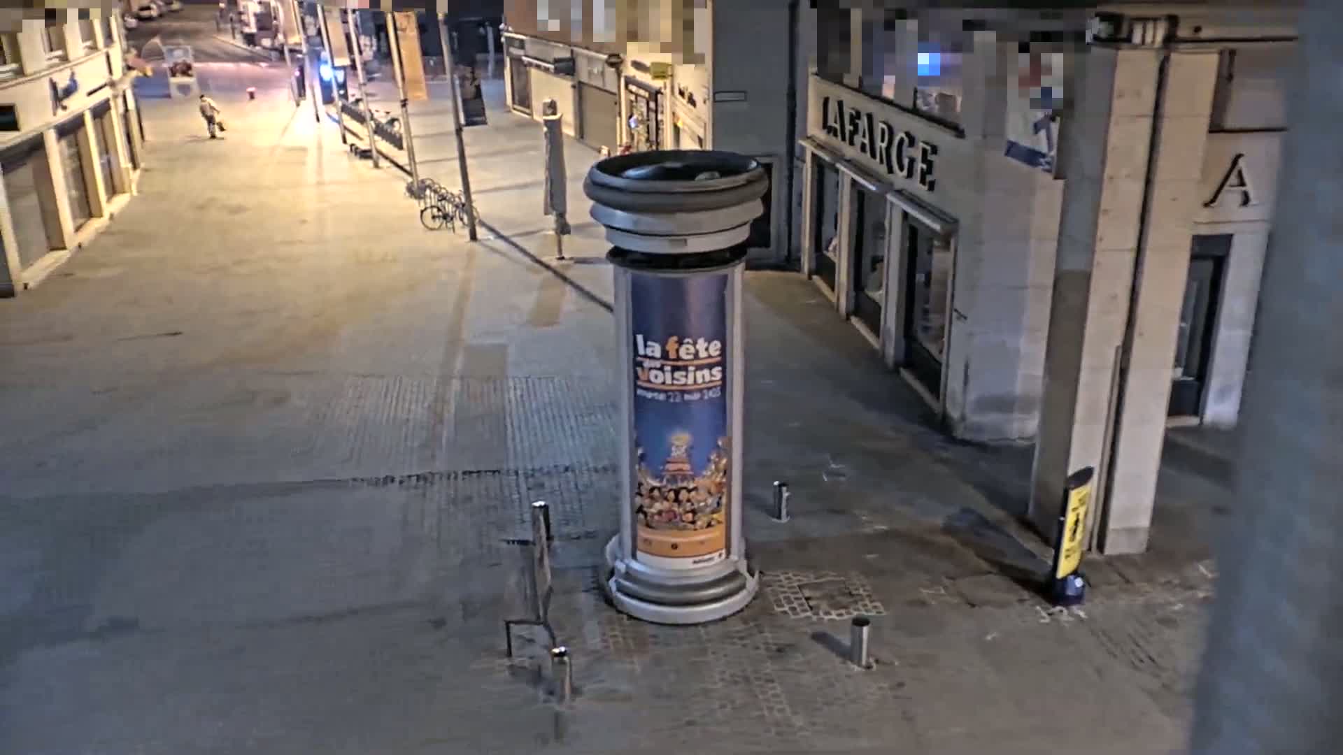 A mostly empty city street at night is seen from an elevated vantage point, showing a cylindrical advertisement stand in the foreground and a few parked bicycles in the distance.
