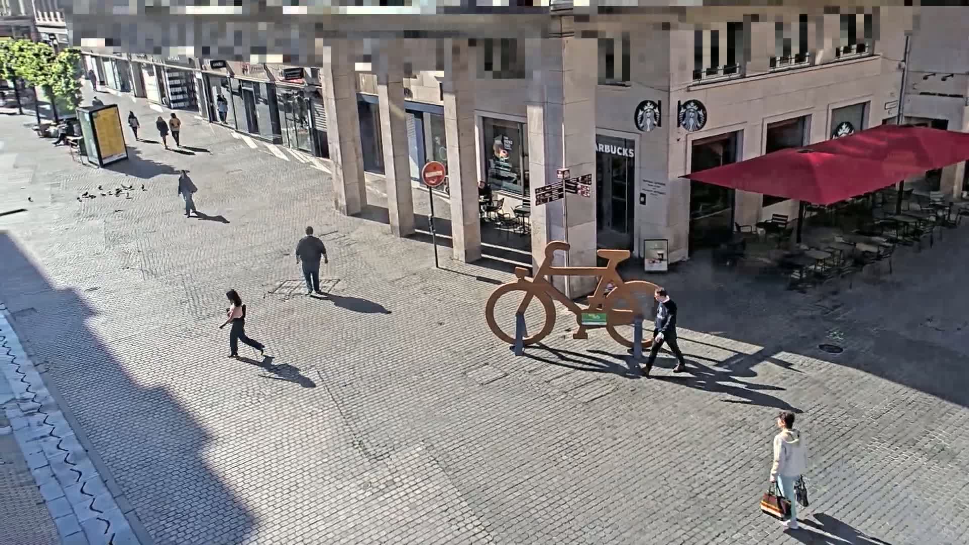 A sunny city street scene shows several pedestrians walking past a Starbucks with outdoor seating and a large,  decorative bicycle sculpture.