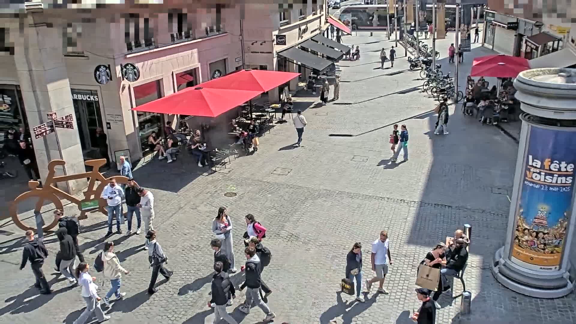 A paved outdoor plaza with people walking, sitting at outdoor cafes under red umbrellas, and a large bicycle sculpture, all under sunny conditions.