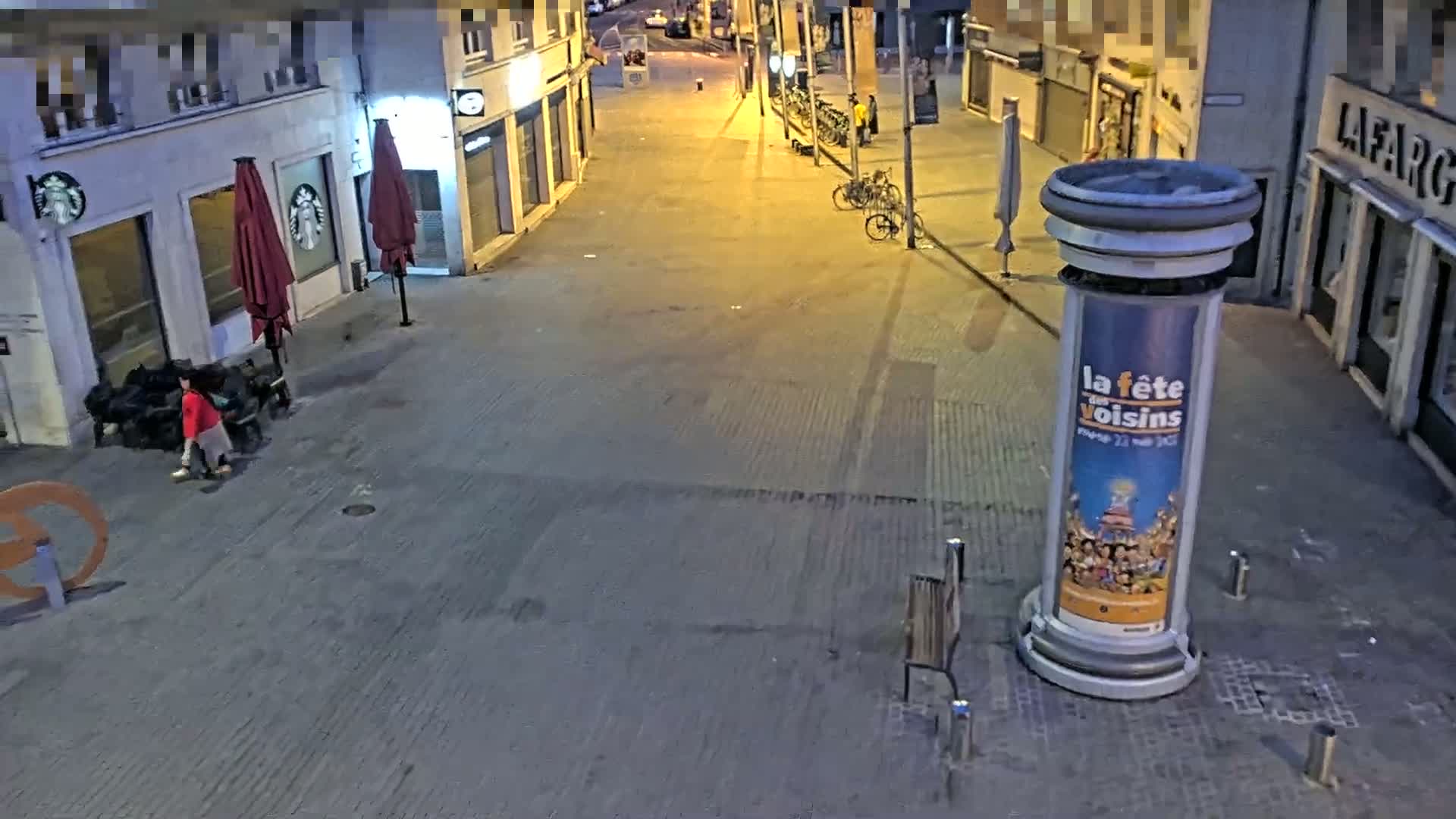 A mostly empty city street at night is lit by streetlights, showing a few people near a Starbucks and some parked bicycles.