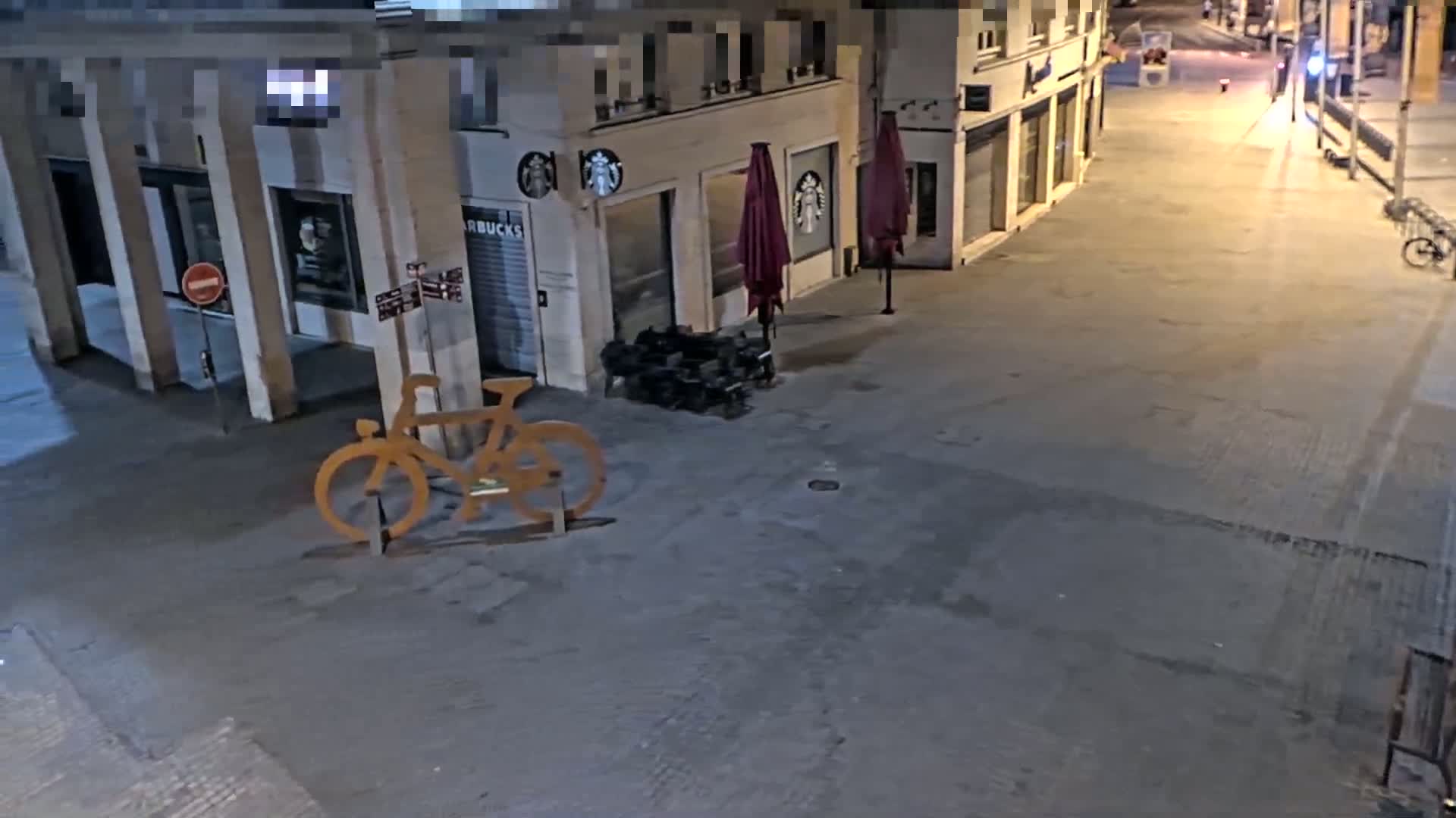 A large, light-colored, wooden bicycle sculpture sits on a nighttime street in front of a building with closed storefronts and maroon umbrellas.