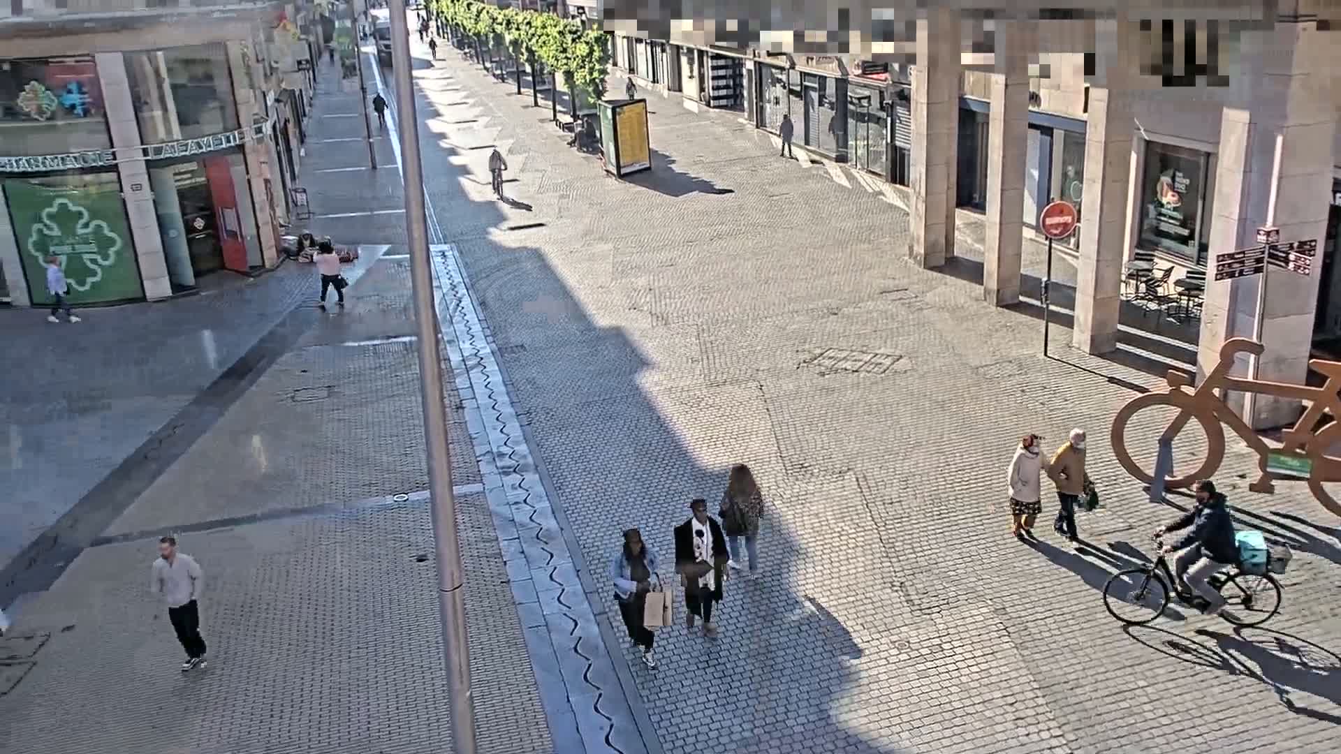 A mostly empty cobblestone city street, under sunny conditions, features a few pedestrians and cyclists, with buildings lining both sides.