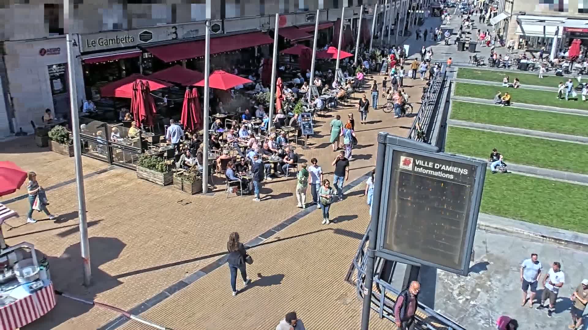 A sunny outdoor plaza scene shows numerous people sitting at cafe tables under red umbrellas and others walking around, with some people relaxing on grassy areas.