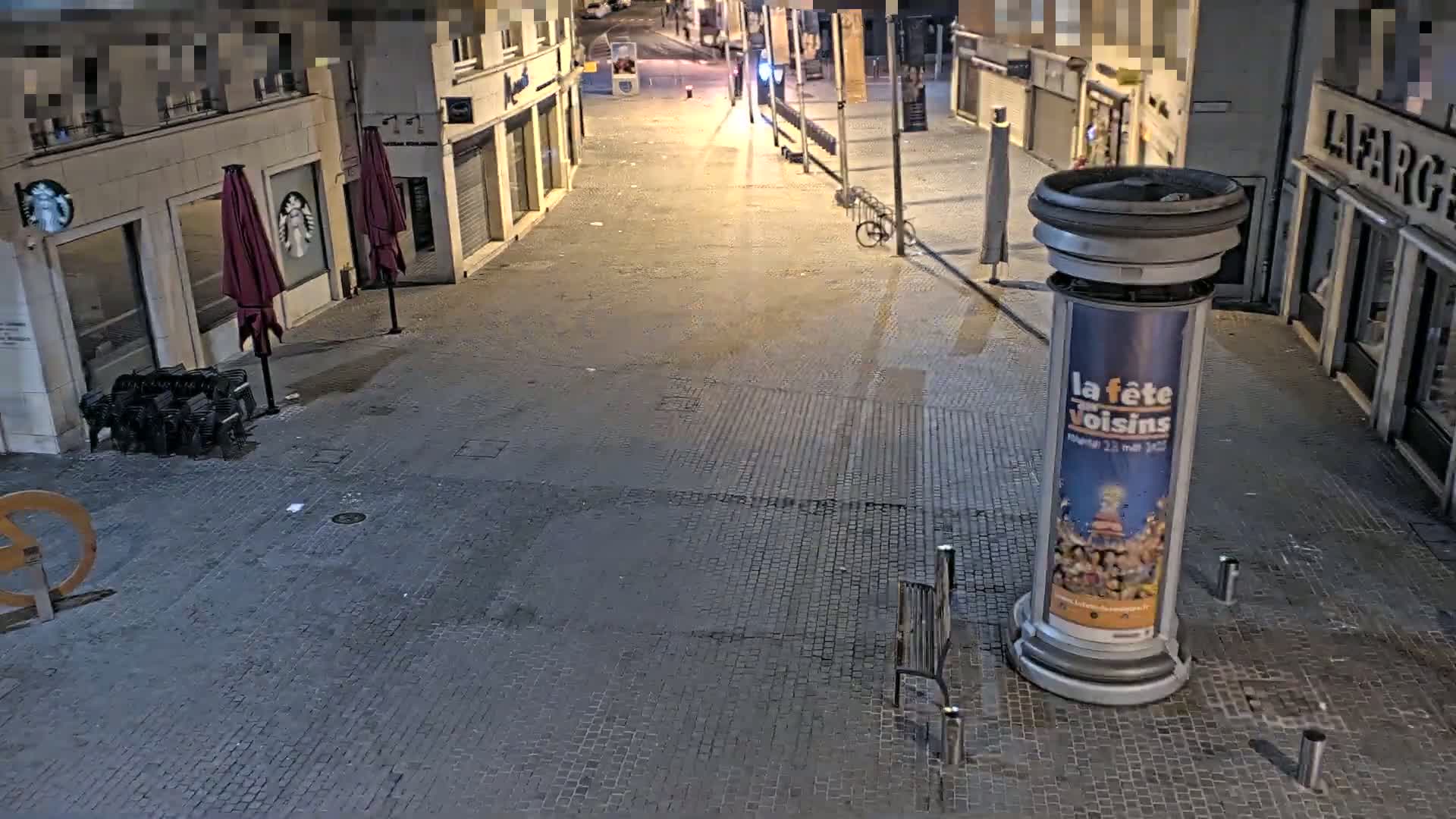 A mostly empty cobblestone street at night is lit by streetlights, with a few chairs and a bicycle visible.