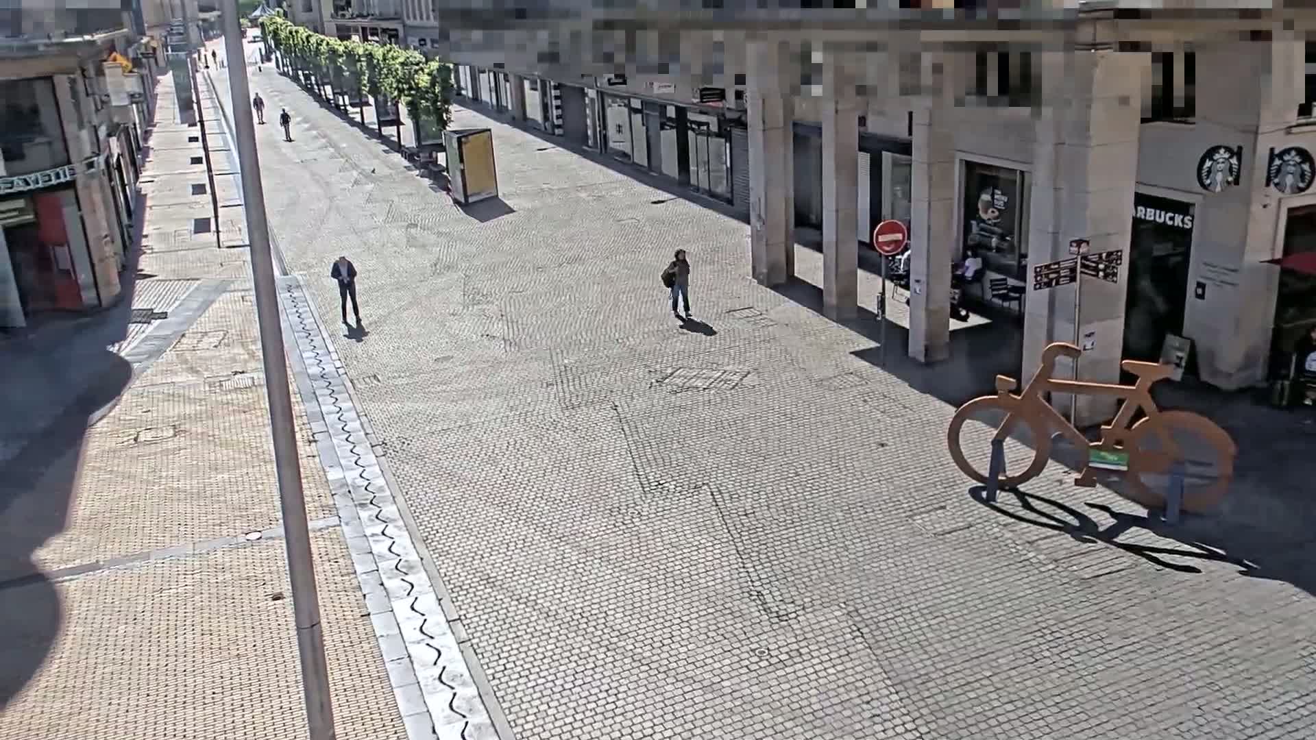 A mostly empty cobblestone street lined with buildings, under sunny conditions, features a few pedestrians and a large bicycle sculpture.