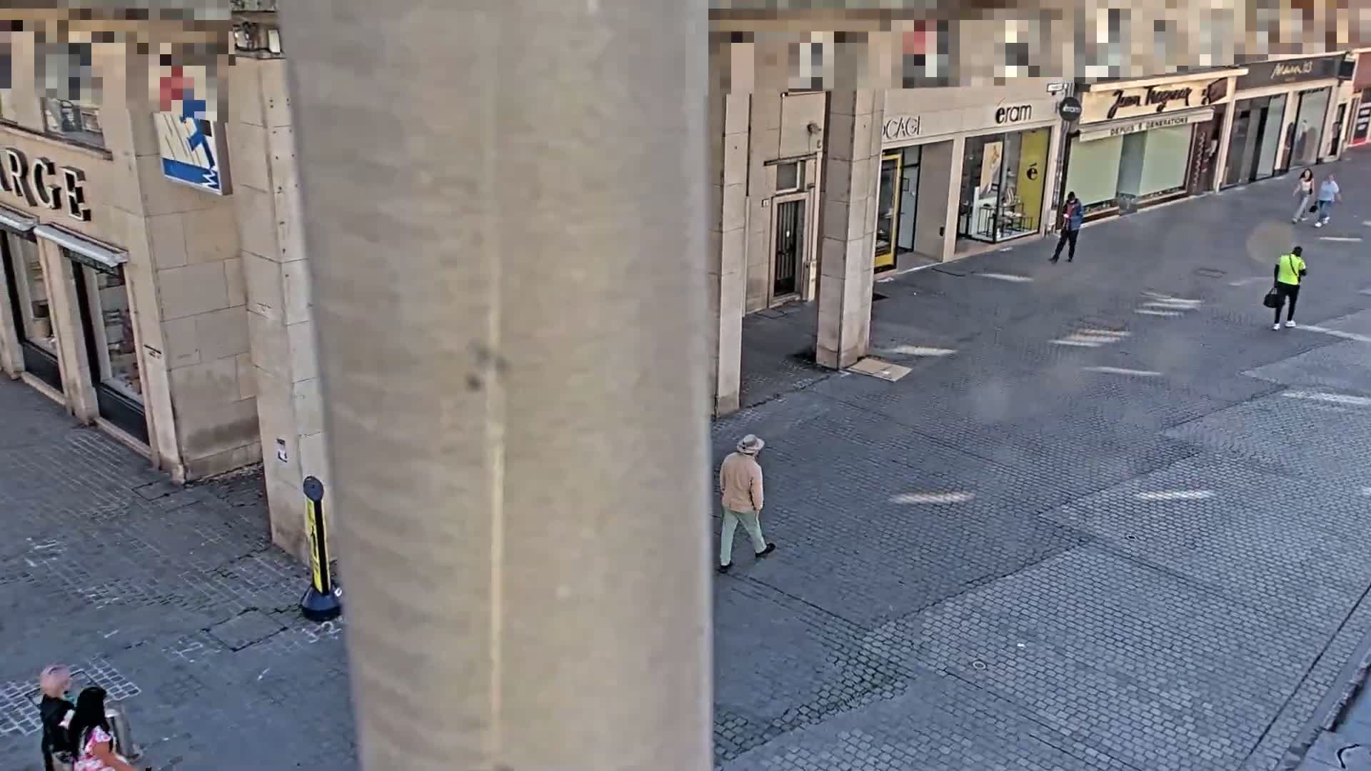 A high-angle view shows a mostly empty cobblestone street with several pedestrians walking past storefronts on a sunny day.